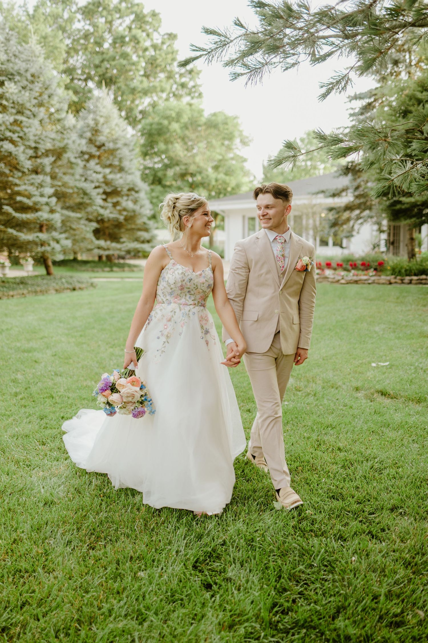 A couple walks hand in hand on a garden lawn, with the bride in white and groom in beige.