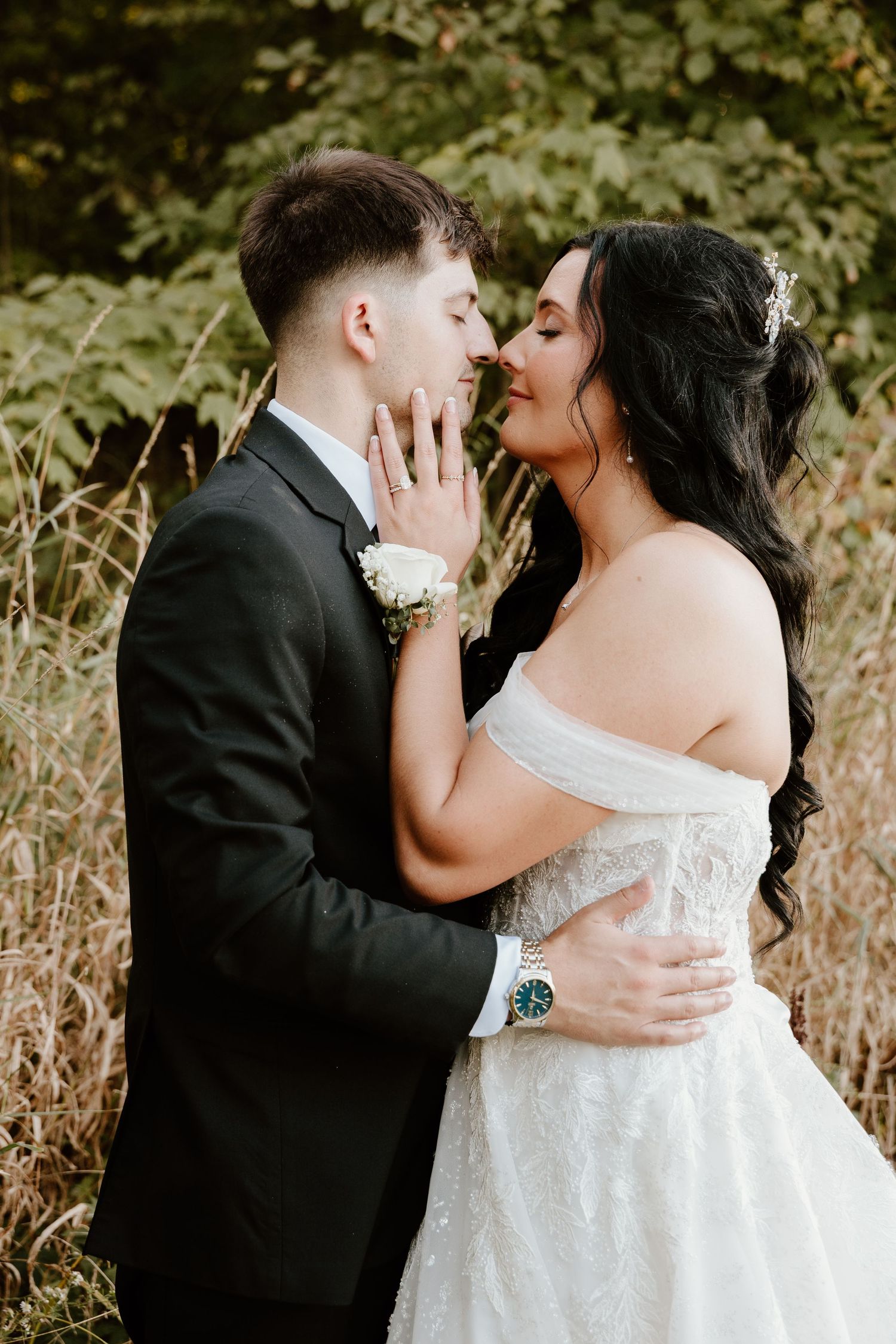 A romantic wedding couple embraces in a sunlit outdoor setting with natural foliage in the background.