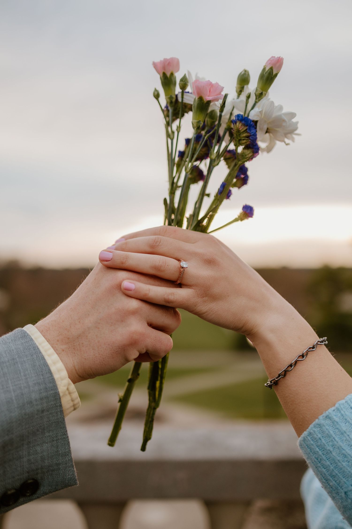 Two hands exchange a small bouquet of flowers with white daisies and blue blooms against a blurred background.