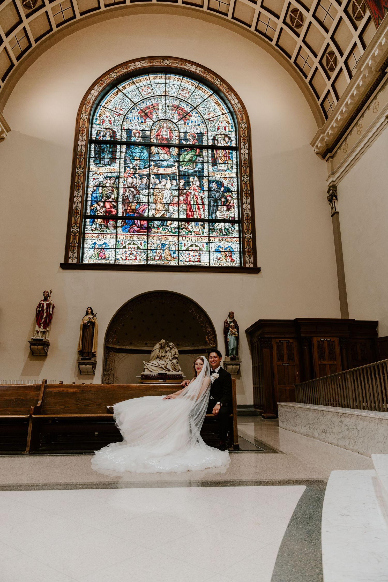 Bride and groom pose before stunning stained glass window in elegant church during wedding ceremony.