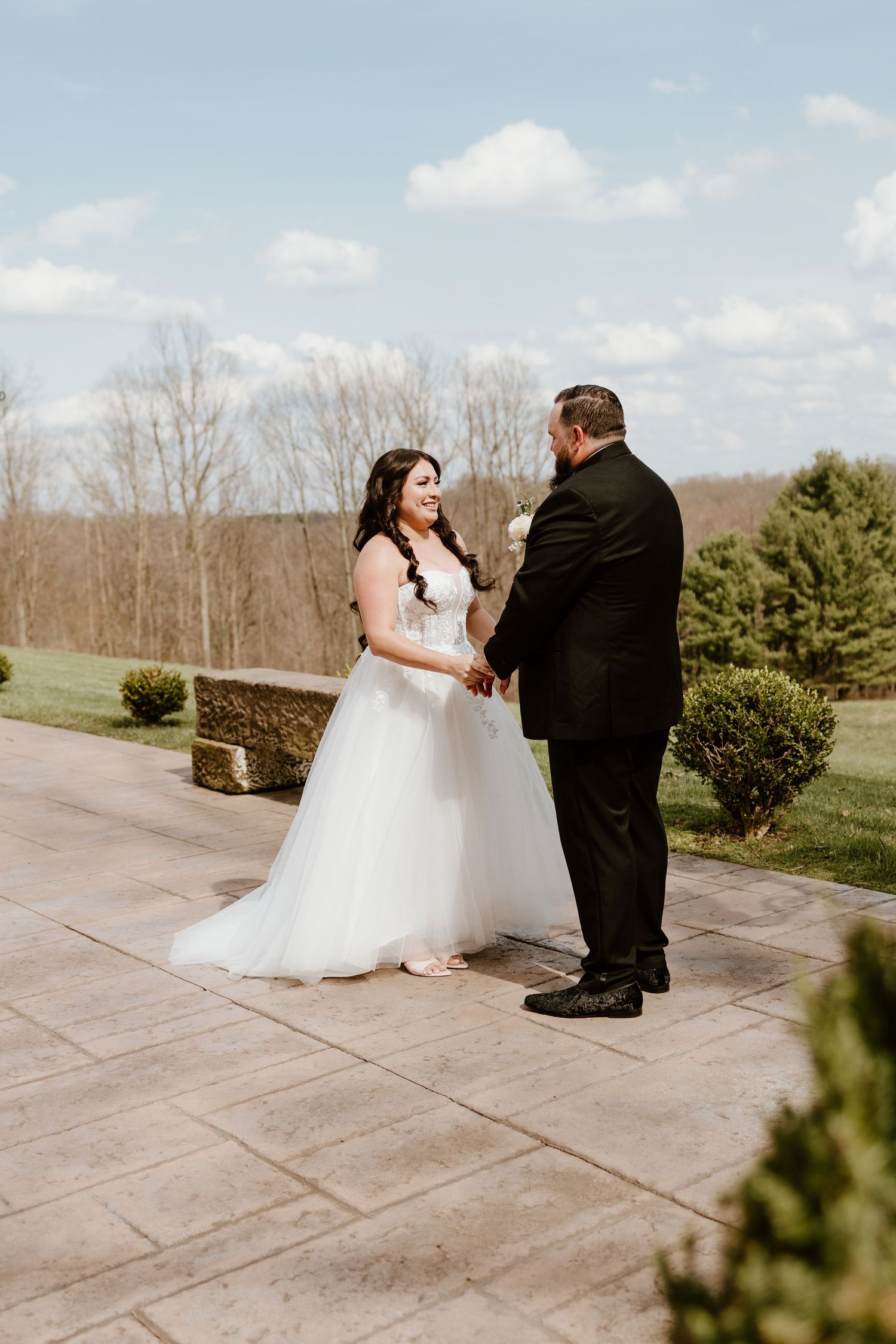 Bride and groom hold hands outdoors on a stone patio, surrounded by lush greenery under a bright blue sky.