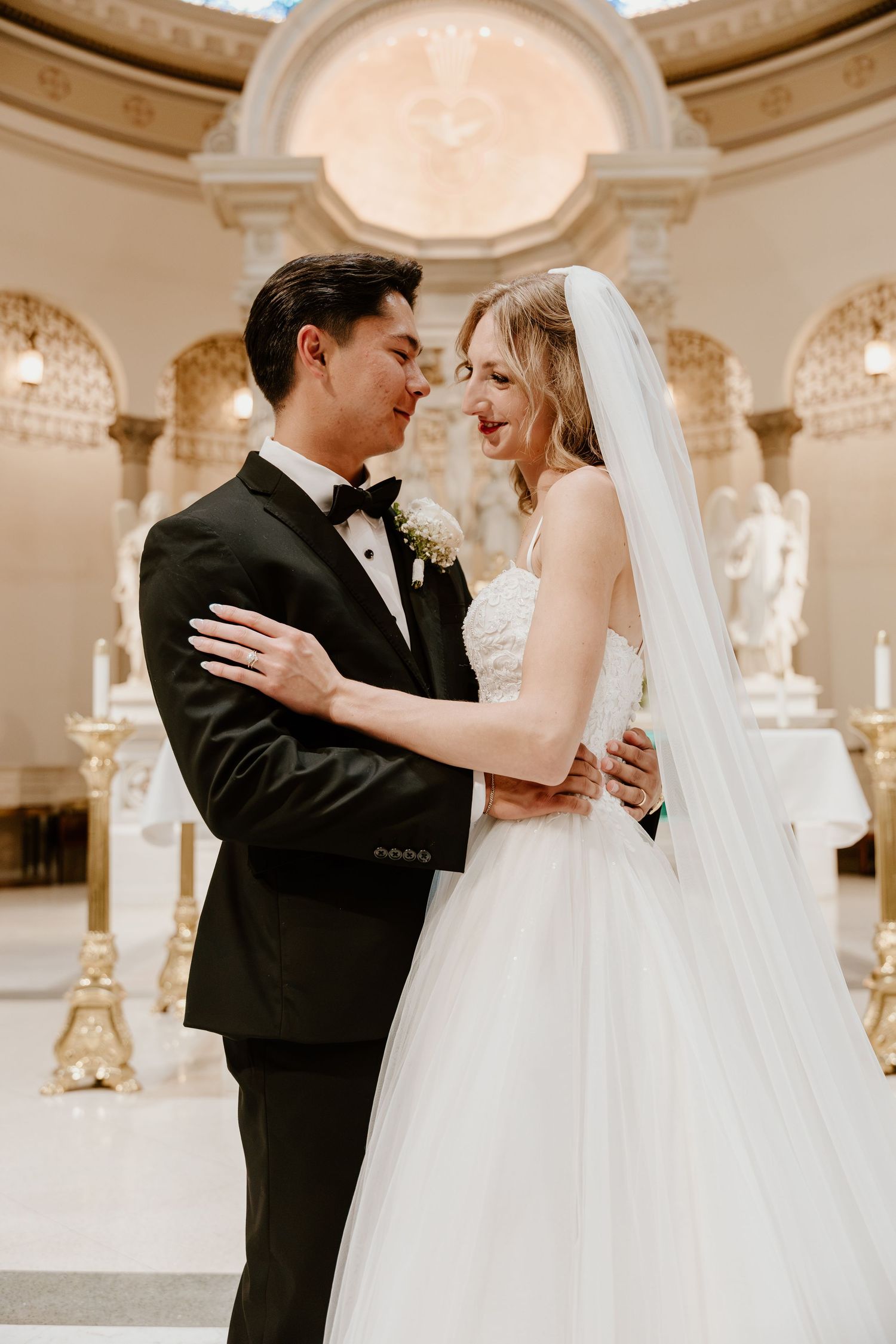 A bride and groom share a romantic moment in an elegant church, she in a white tulle gown with veil, he in a black tuxedo.