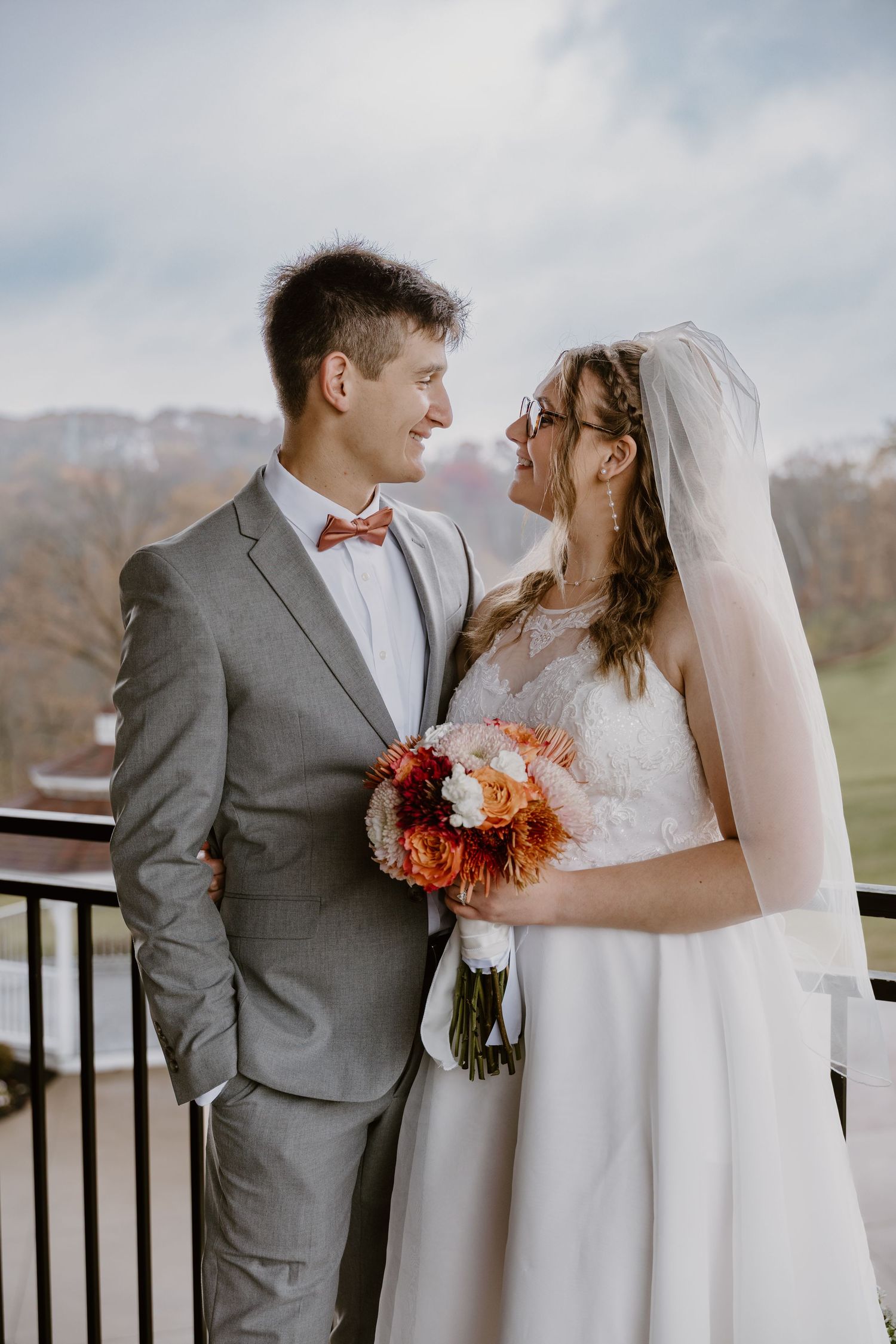 A bride and groom share a romantic moment on a balcony, she holds an orange bouquet, wearing a veil and white gown.