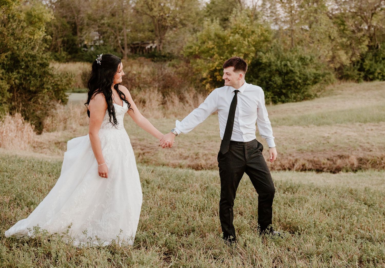 A bride and groom hold hands dancing in a green field on their wedding day.