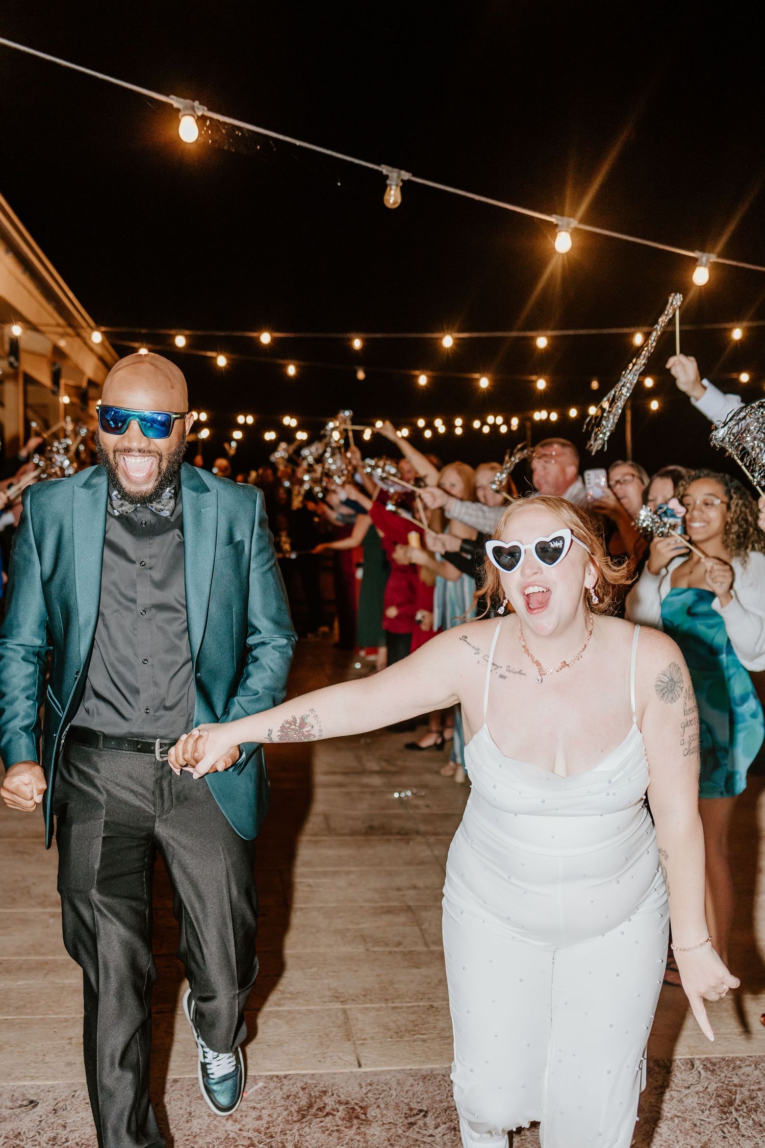 Joyful newlyweds in sunglasses walk through sparkler exit at night wedding reception with string lights.