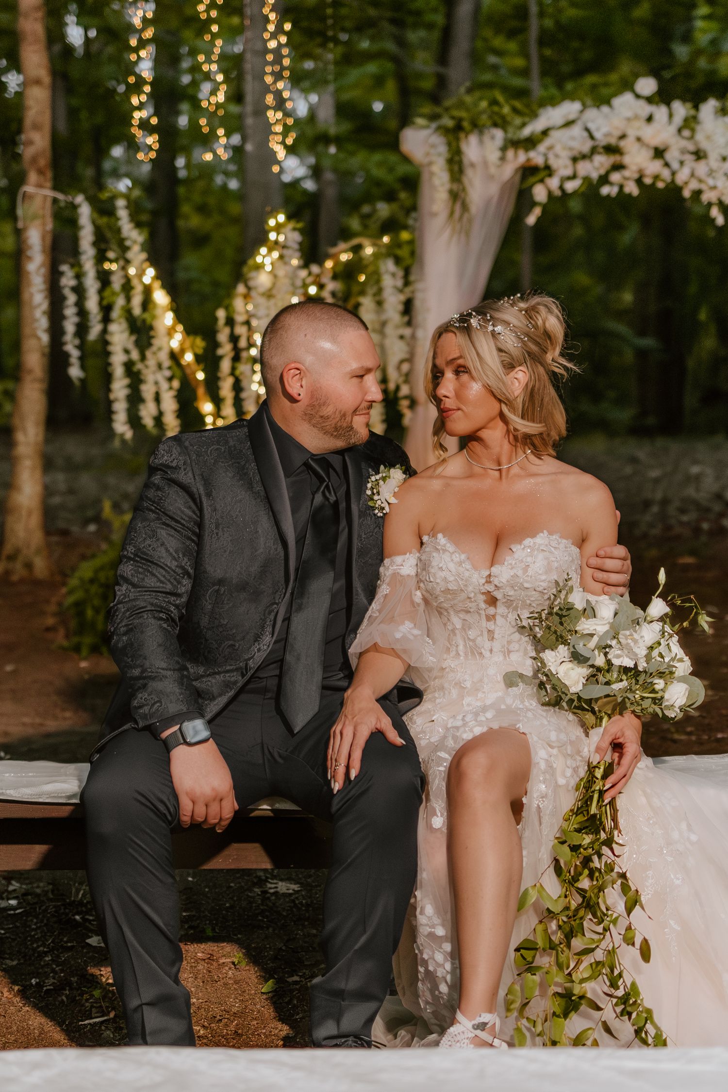 Romantic wedding couple shares intimate moments on rustic bench surrounded by trees with twinkling lights.