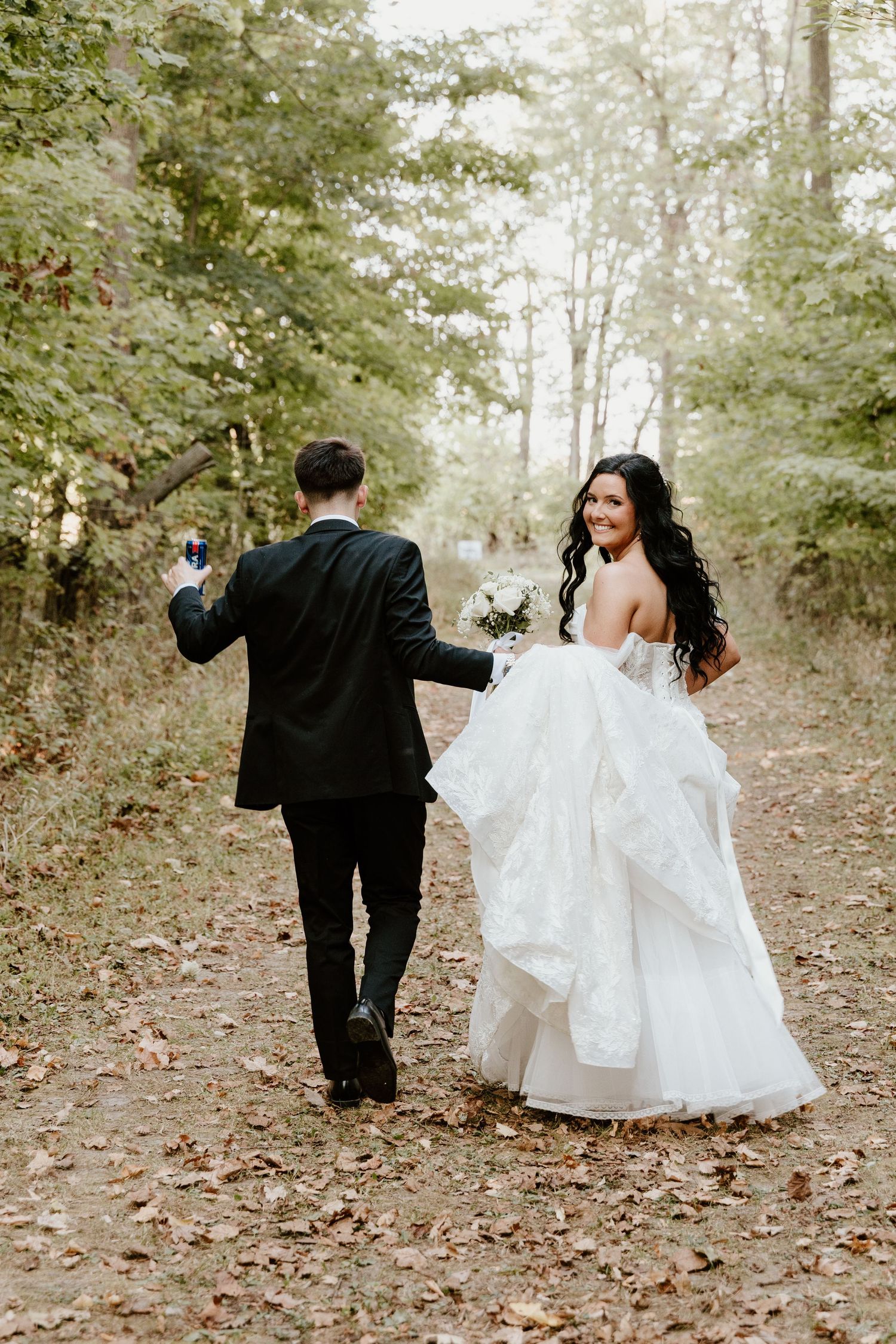 A couple in wedding attire walks down a leaf-covered forest path while holding hands on their special day.