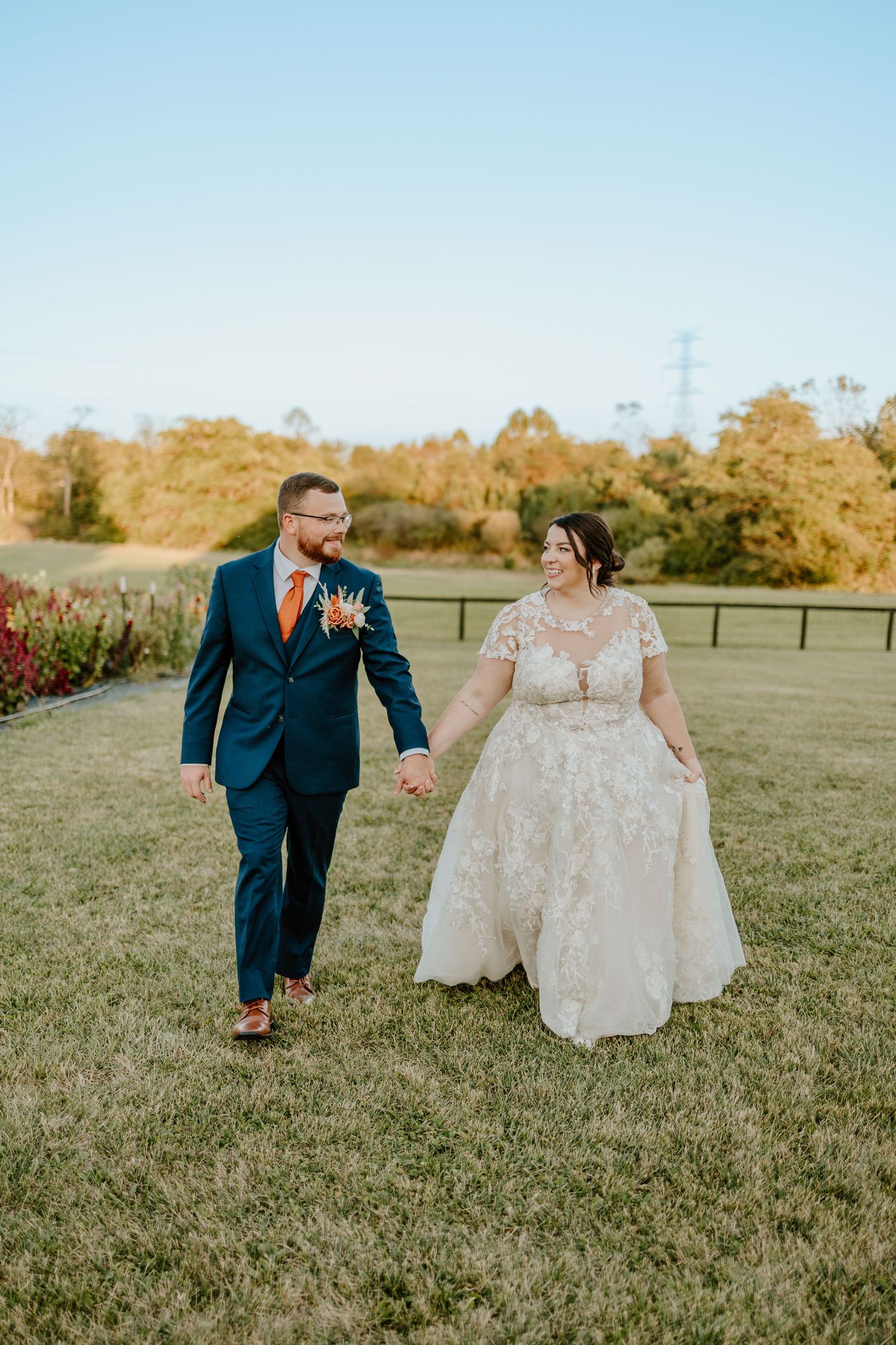 A couple walks together on a grassy field during golden hour, with the bride wearing a lace gown and groom in navy suit.