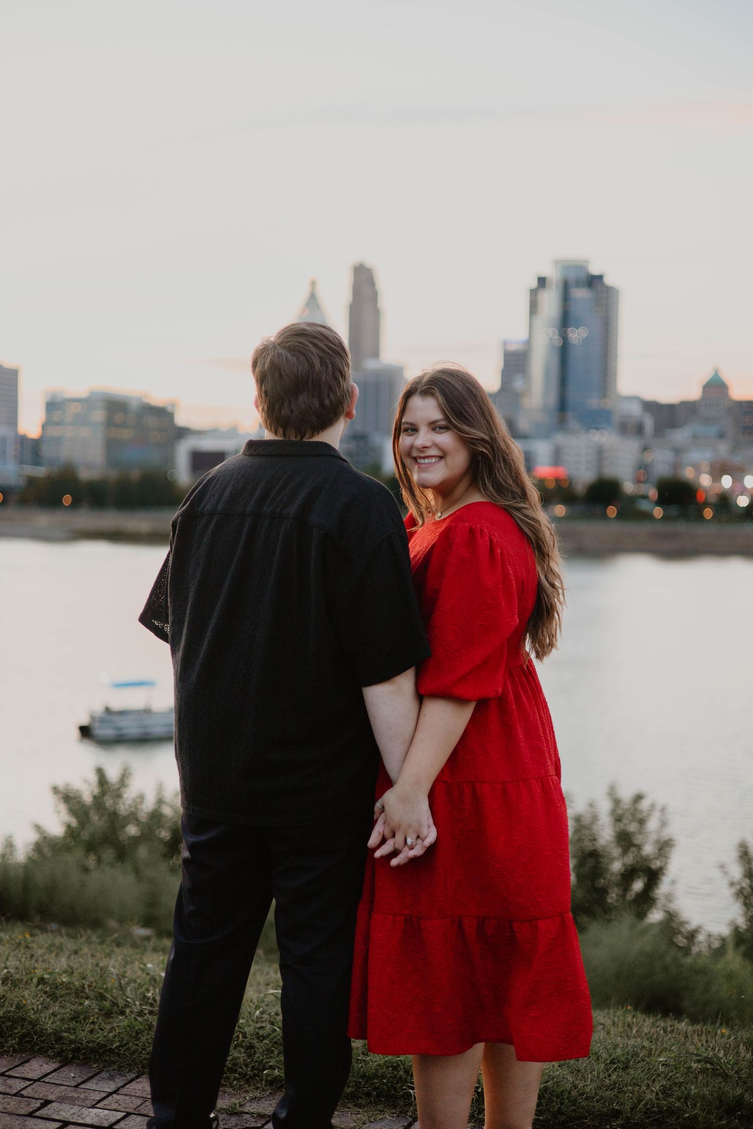 Couple in red dress and black attire embracing by waterfront with city skyline at sunset.