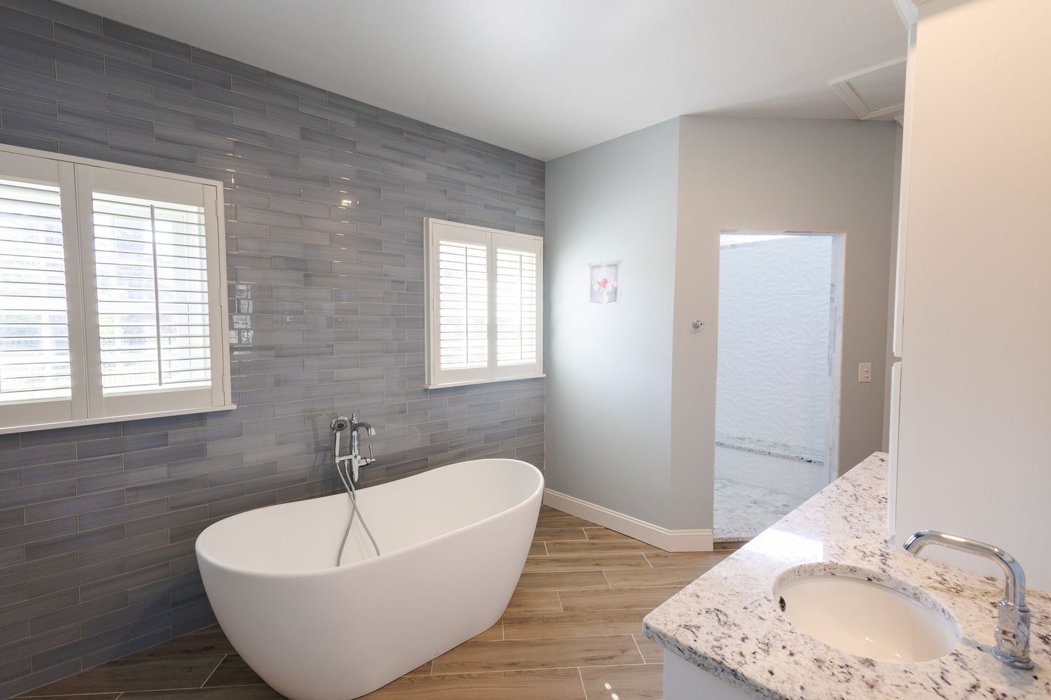 Modern bathroom with freestanding soaking tub, gray stone accent wall, hardwood floors, and marble vanity countertop.