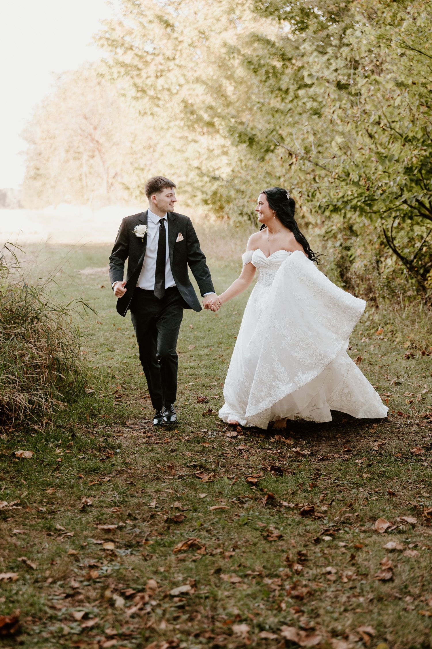 A couple in wedding attire walks hand in hand along a leaf-strewn path surrounded by autumn foliage.