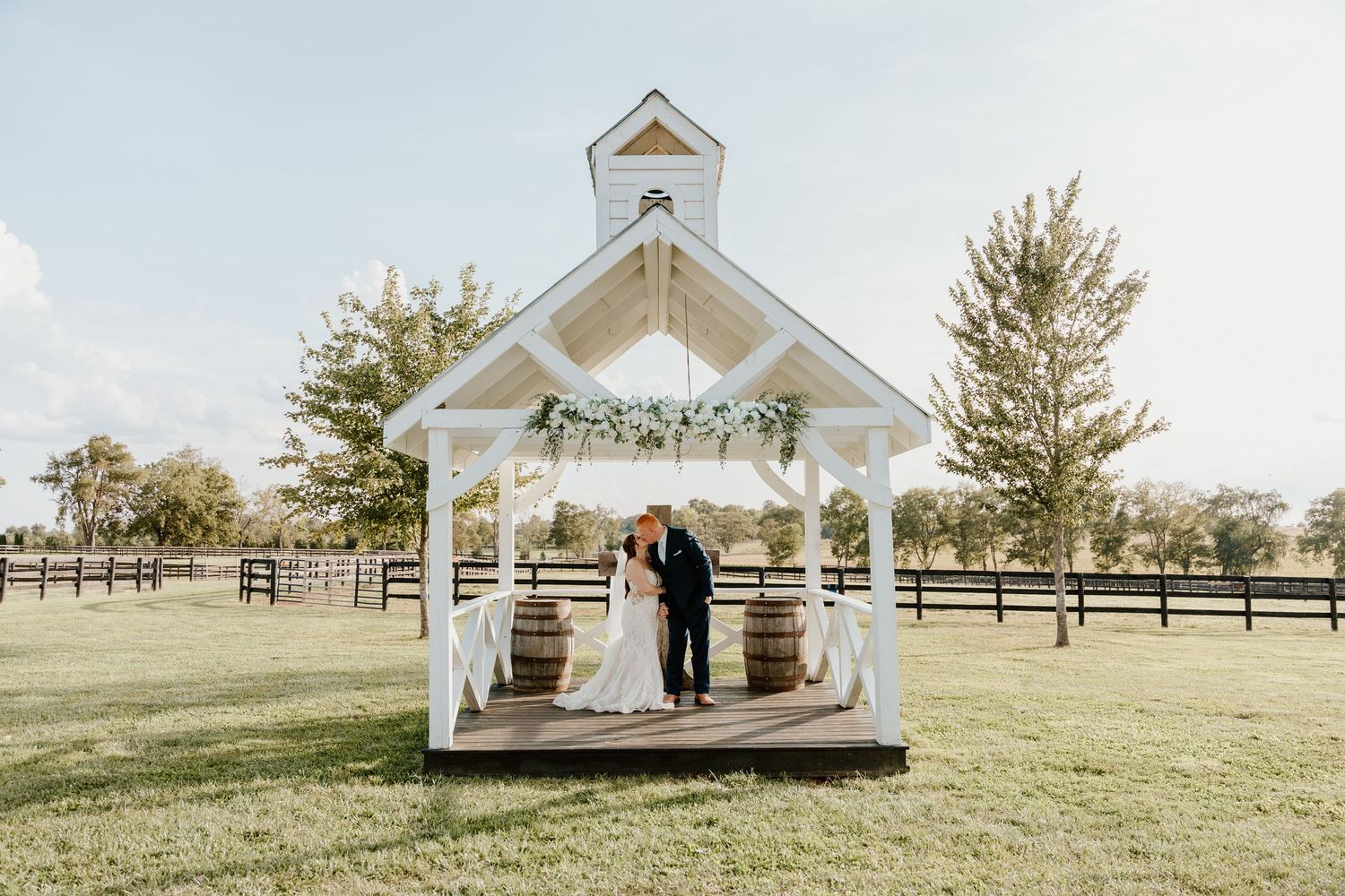 Couple exchanging vows under a white open-air chapel gazebo on a lush green farm with wooden barrel accents.