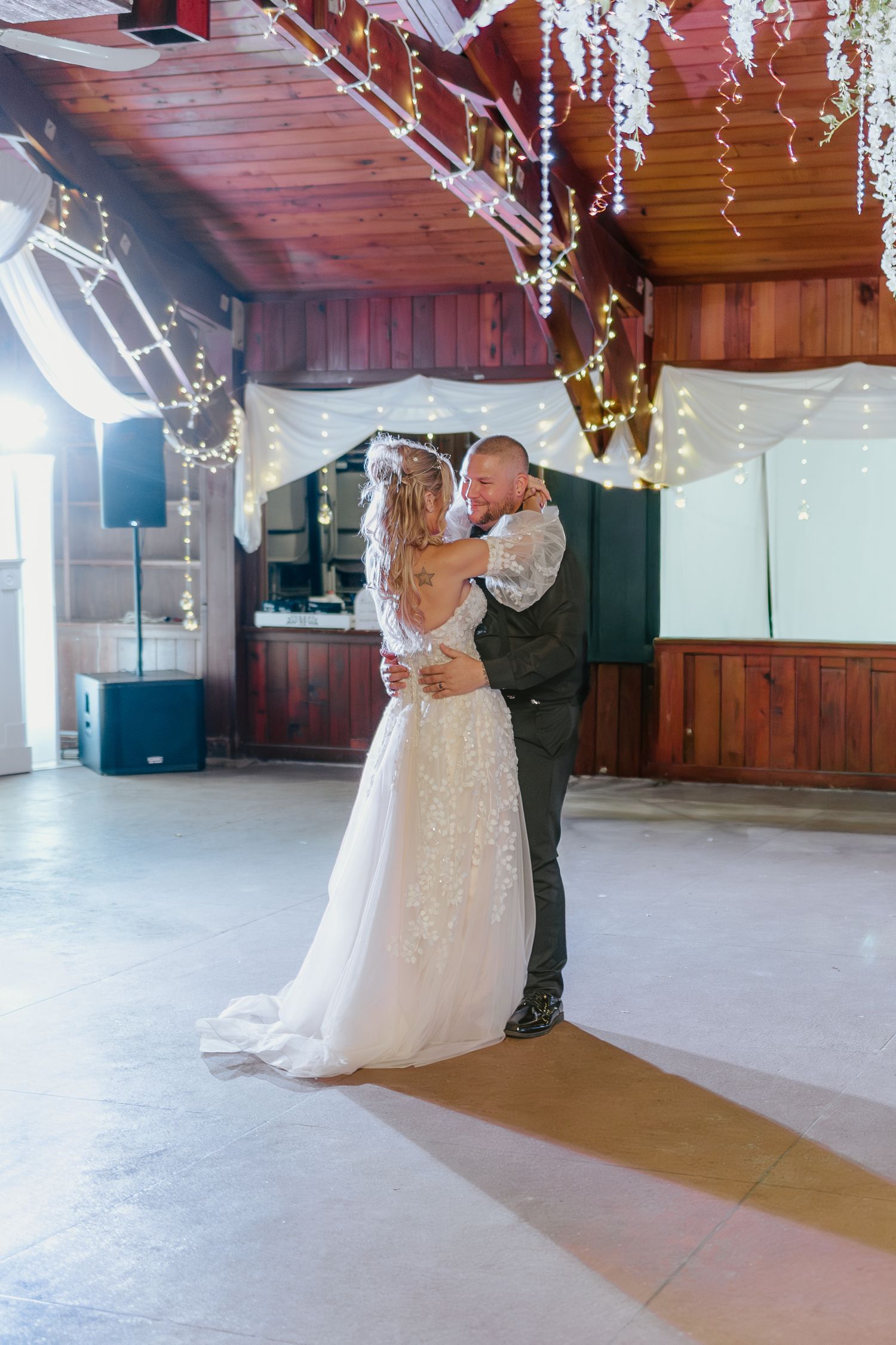 A couple shares their first dance as newlyweds under twinkling lights in a rustic wooden venue.