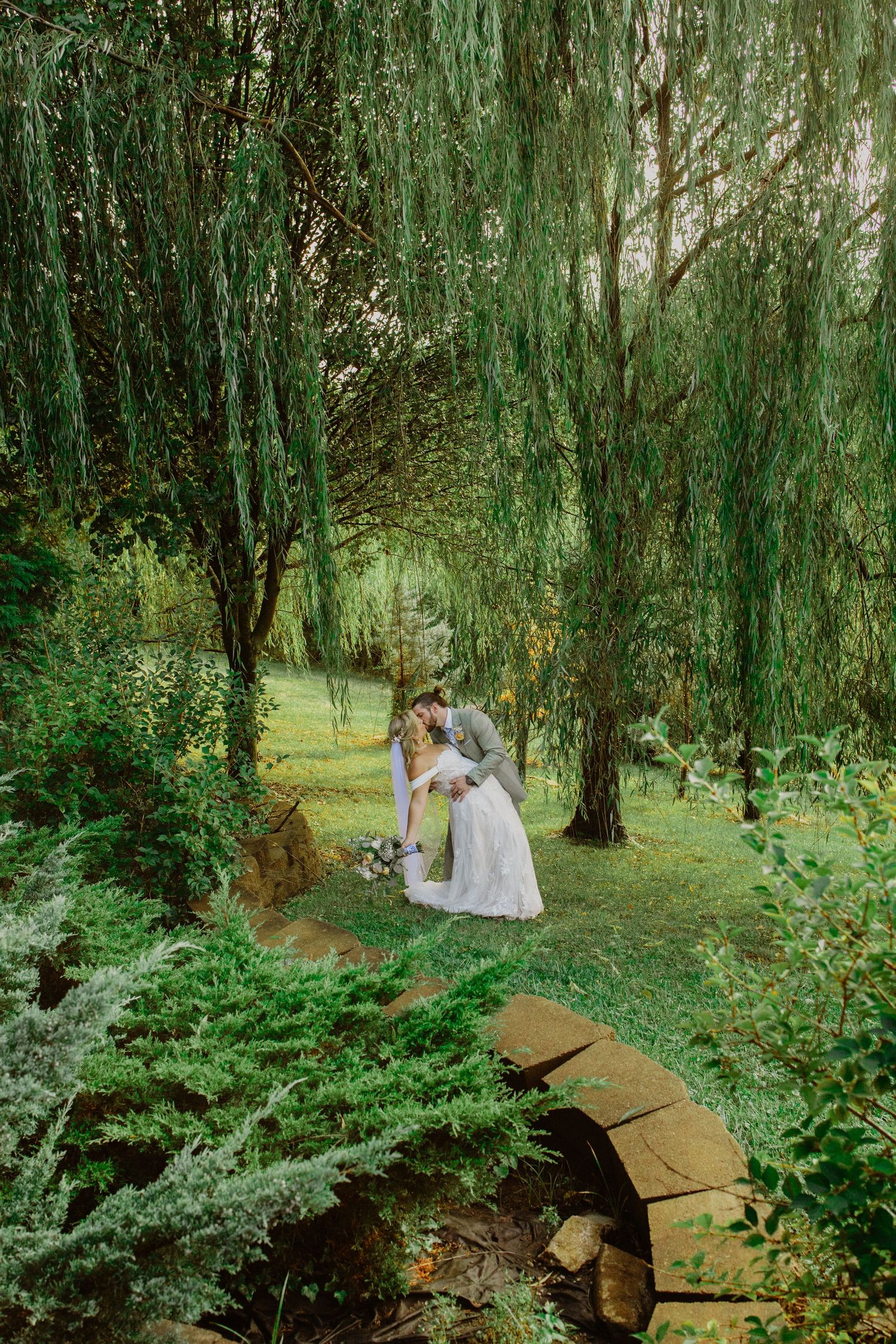 Couple embraces under cascading willow trees in a lush green garden setting during a romantic outdoor photoshoot.