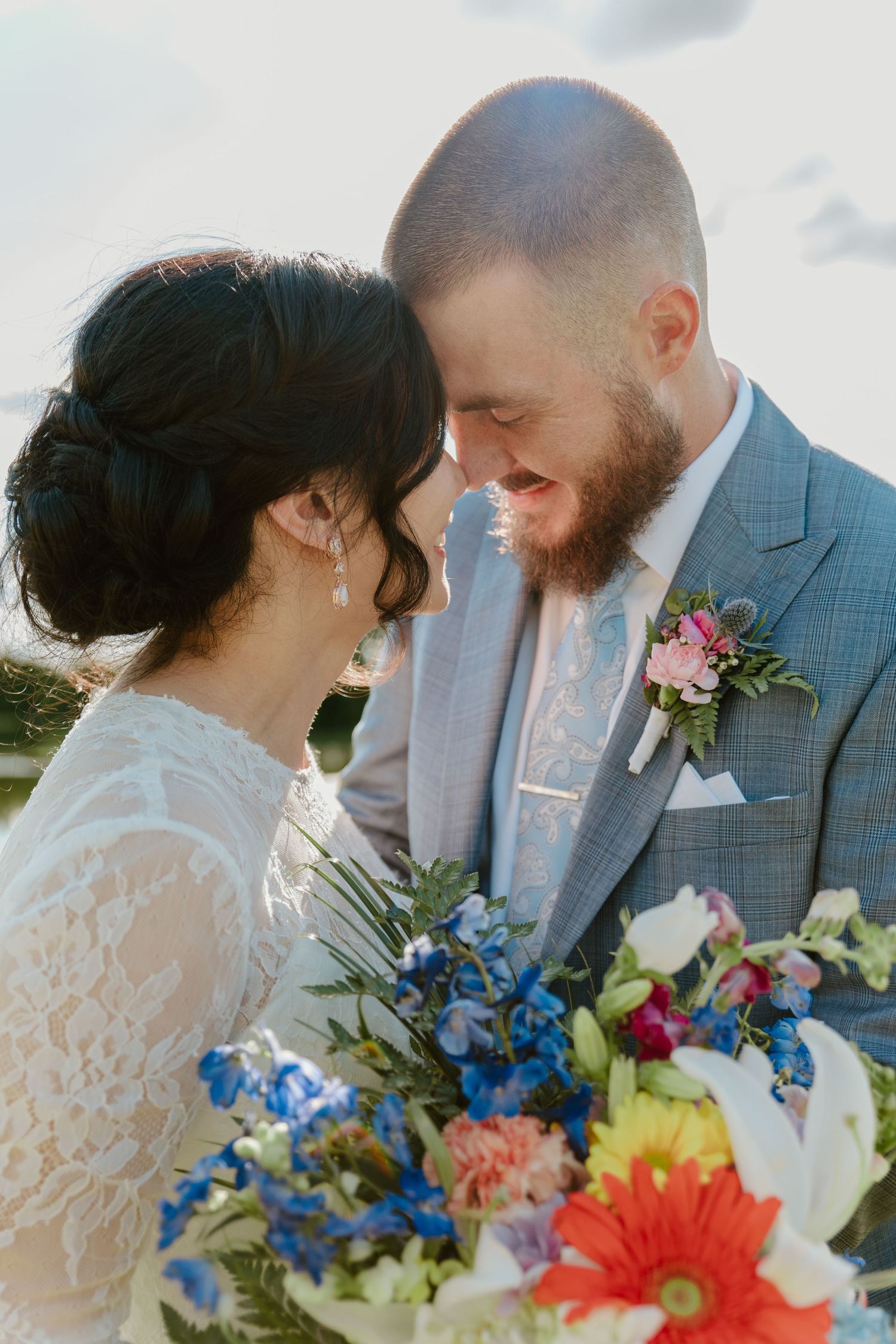 Wedding couple sharing intimate moments outdoors with colorful floral bouquet featuring blue and coral blooms.