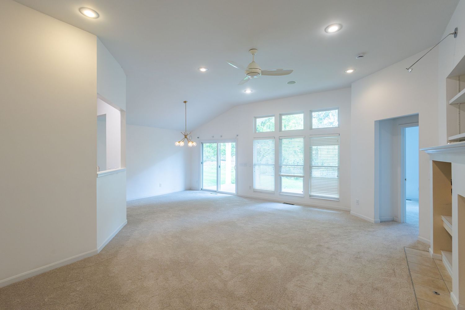 Spacious empty living room with vaulted ceilings, ceiling fan, recessed lighting, large windows, and neutral carpet flooring.