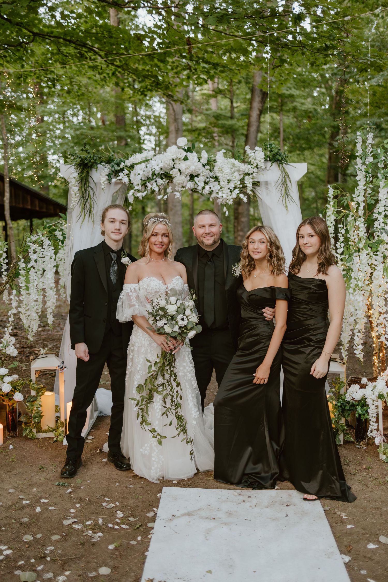 A wedding celebration in a forest setting with white floral decorations and guests in formal attire.