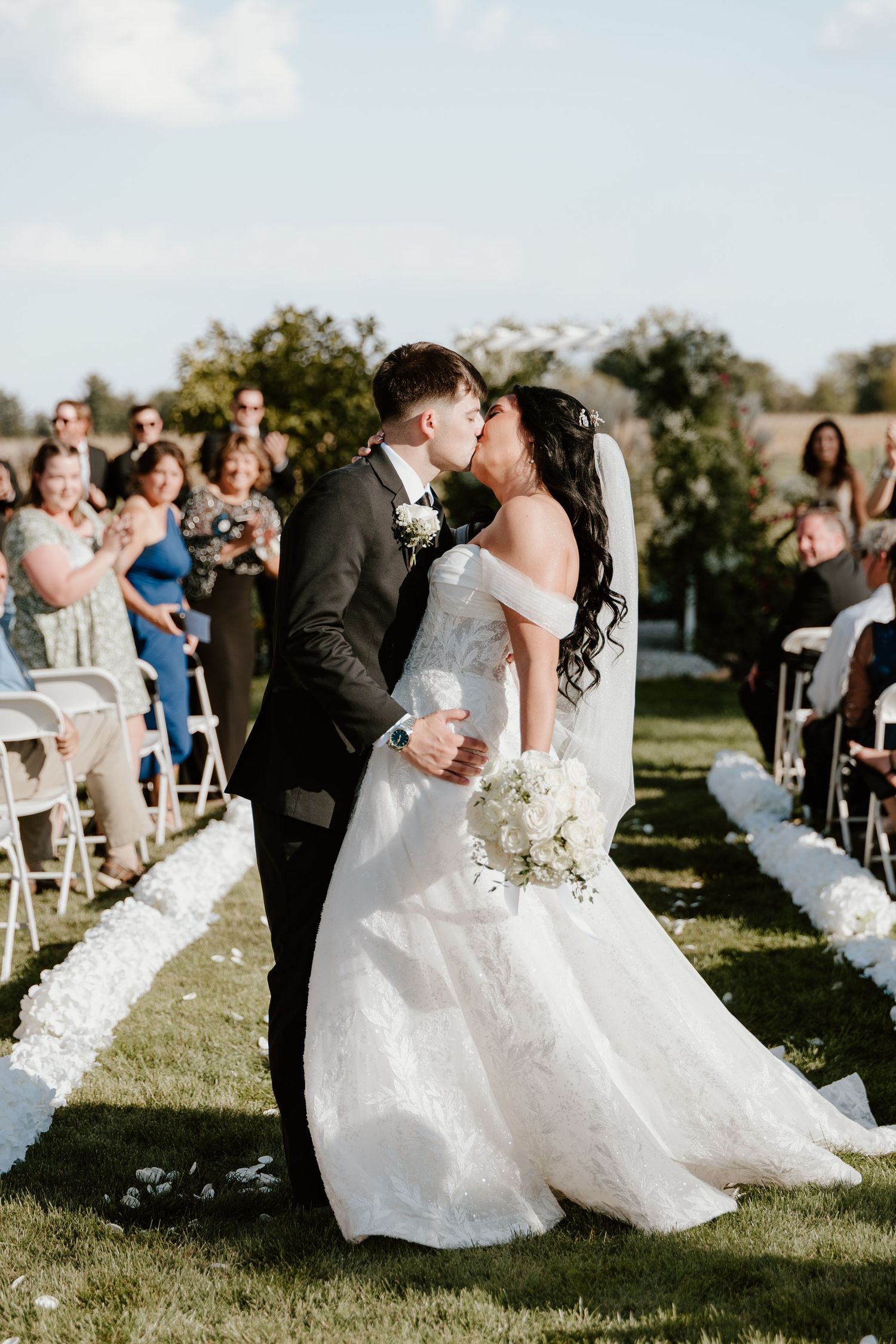 A romantic wedding ceremony takes place outdoors with white chairs and guests watching the couple's first kiss.