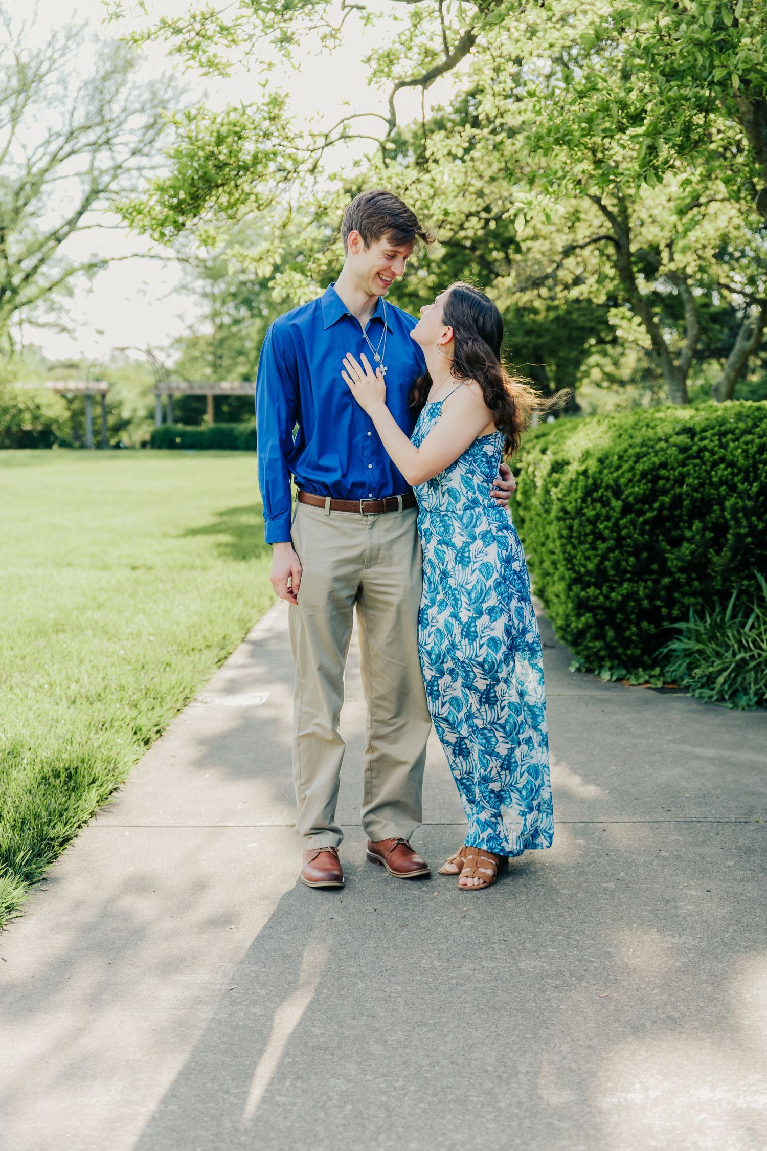 Couple in blue and floral outfits walking and embracing on garden path surrounded by greenery.