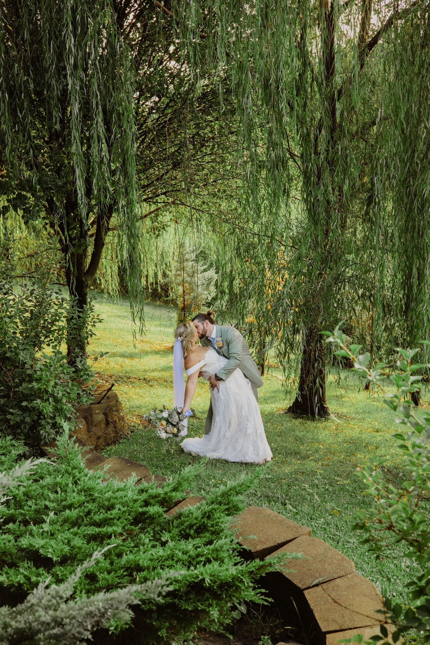 A romantic wedding moment under drooping willow trees with a stone path beneath.