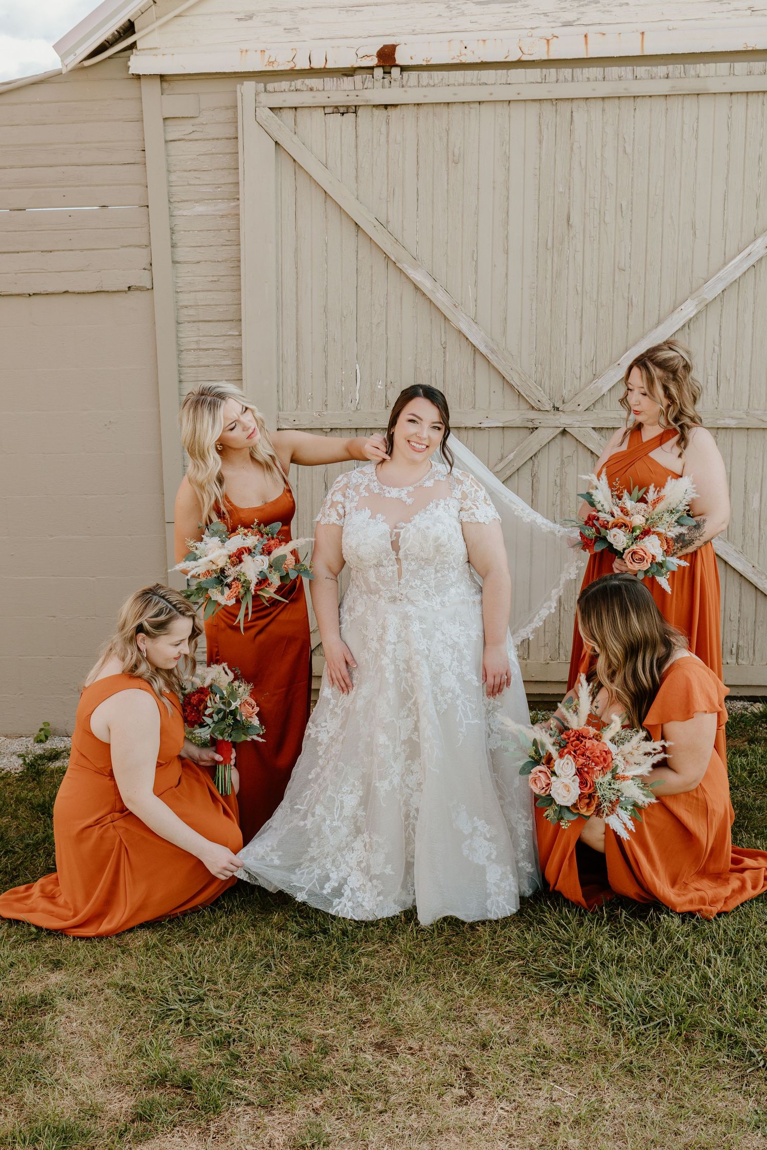 Bride in lace gown surrounded by four bridesmaids in burnt orange dresses holding floral bouquets near a rustic barn.