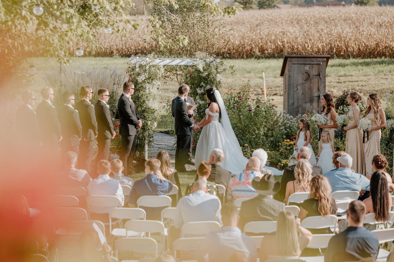 Wedding guests gather outdoors at a scenic venue with white chairs and natural decor for a summer ceremony.