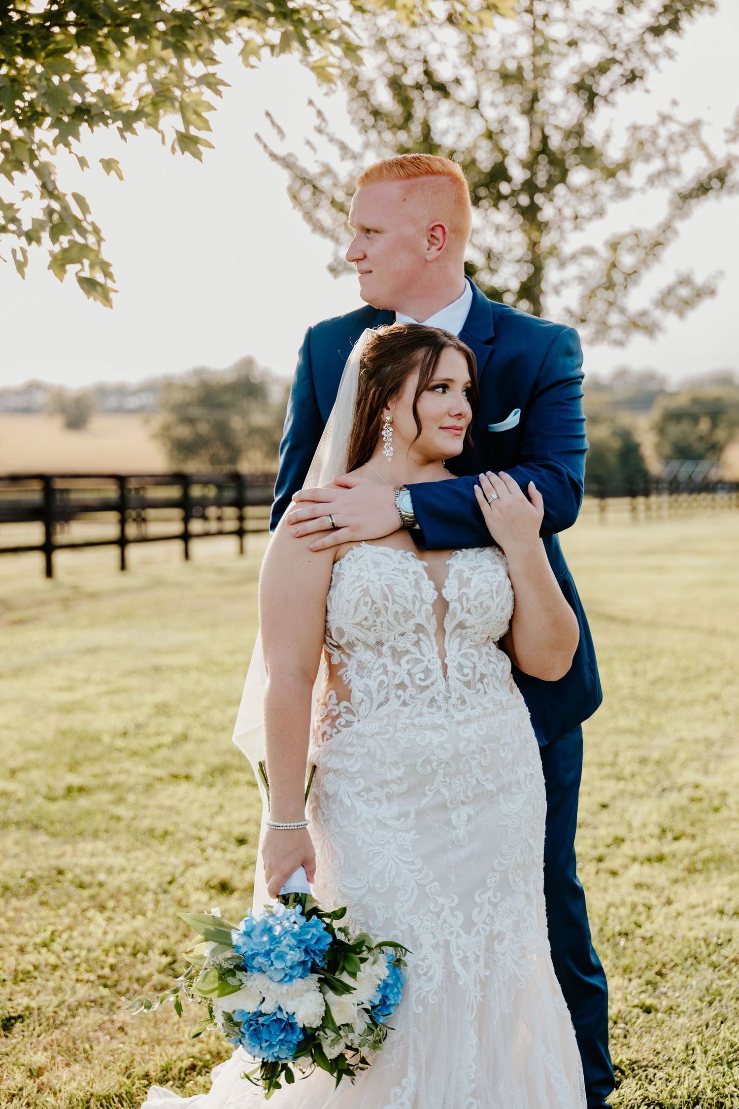 Bride in lace gown holding blue hydrangeas bouquet embraced by groom in navy suit outdoors at golden hour.