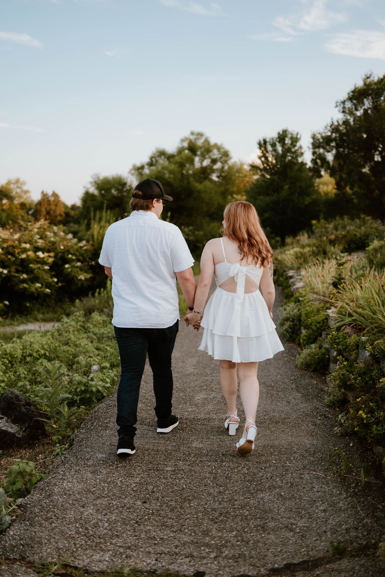 Couple walking hand in hand on a garden path during sunset wearing white and casual attire.