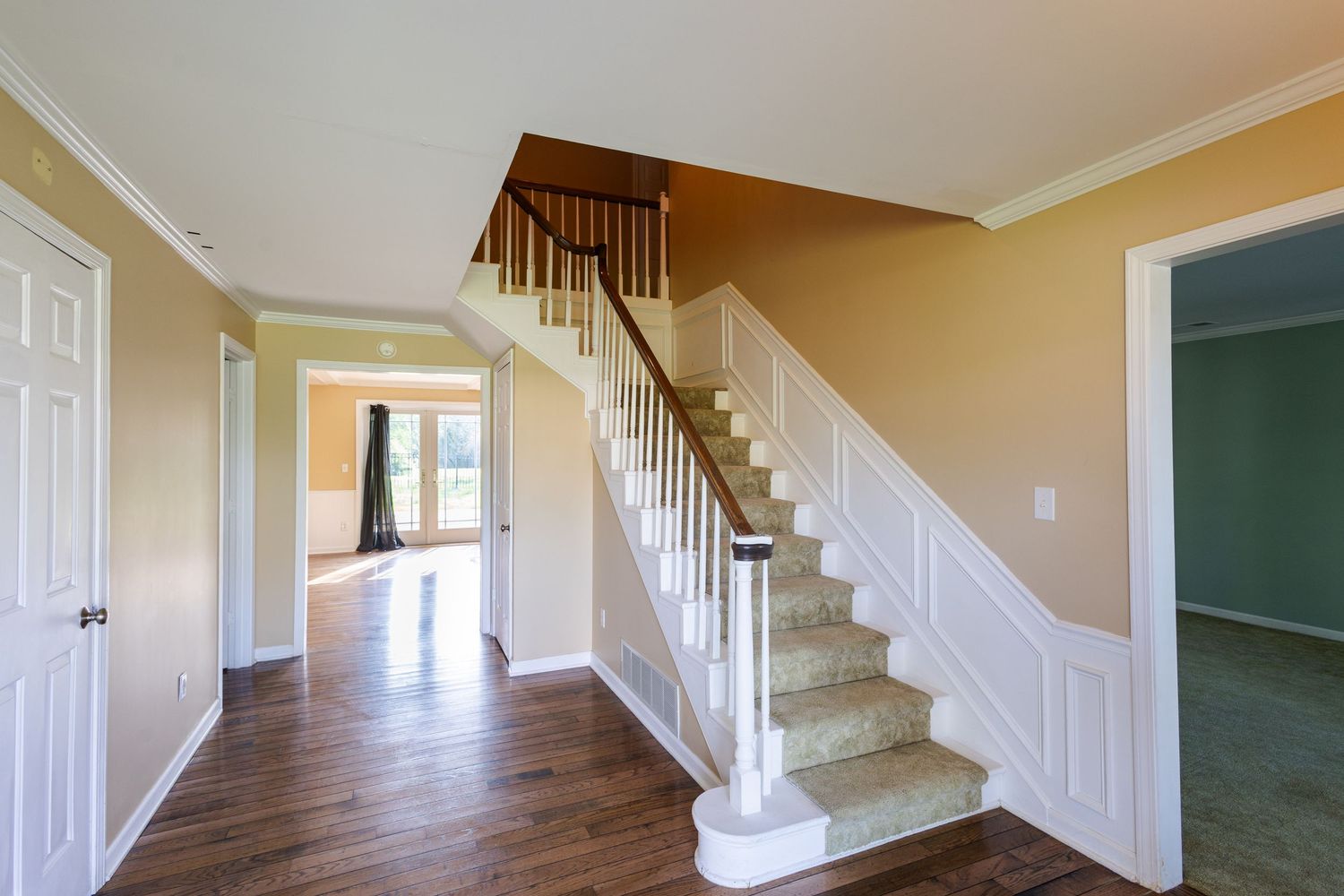 Bright empty foyer with white staircase, hardwood floors, crown molding, and open doorway leading to sunlit room.