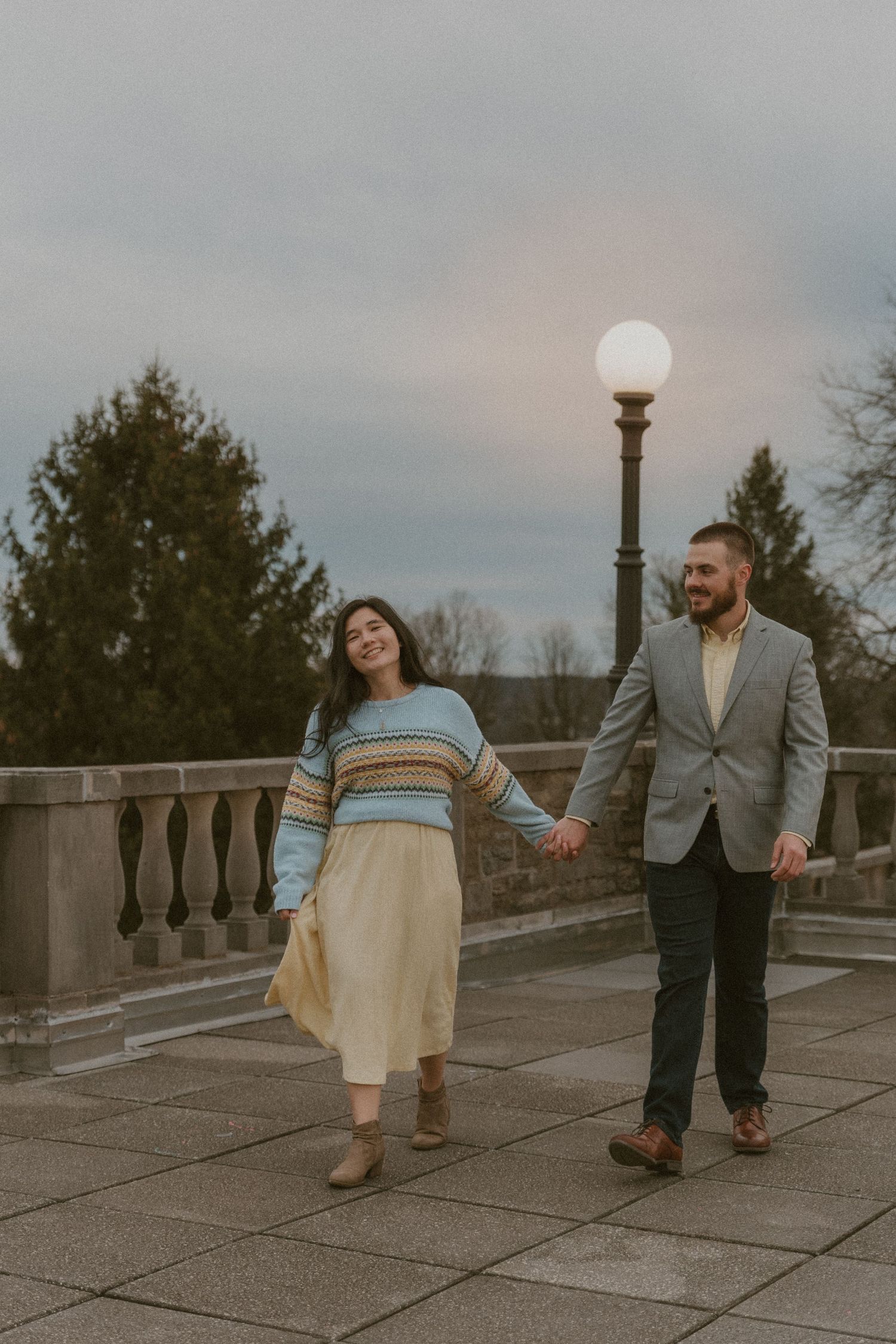 Two people walk together on a concrete terrace with lampposts and trees in the background.