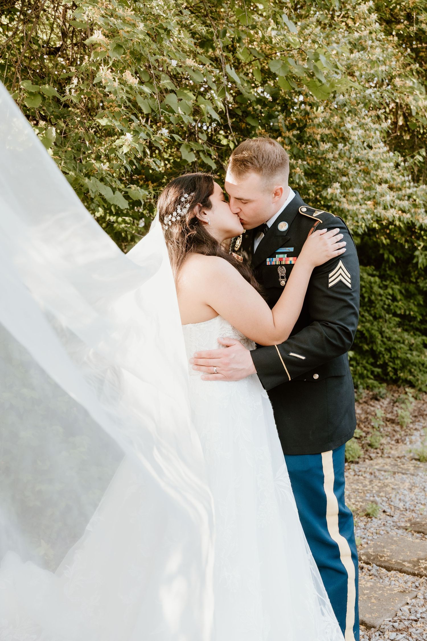 Military groom in dress uniform kissing bride in white gown outdoors, veil flowing in the breeze.