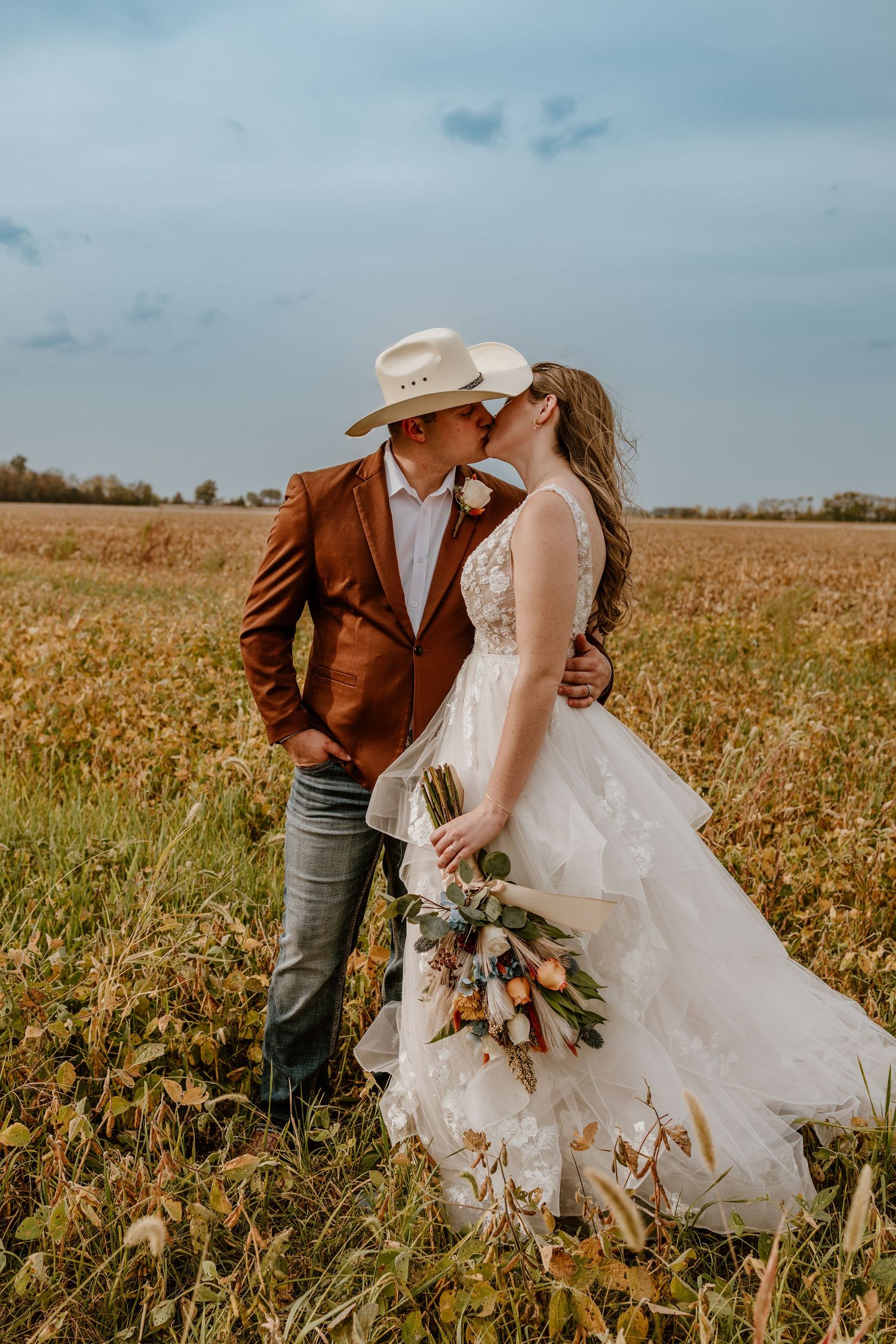 A romantic couple in wedding attire embraces in a golden autumn field during sunset.