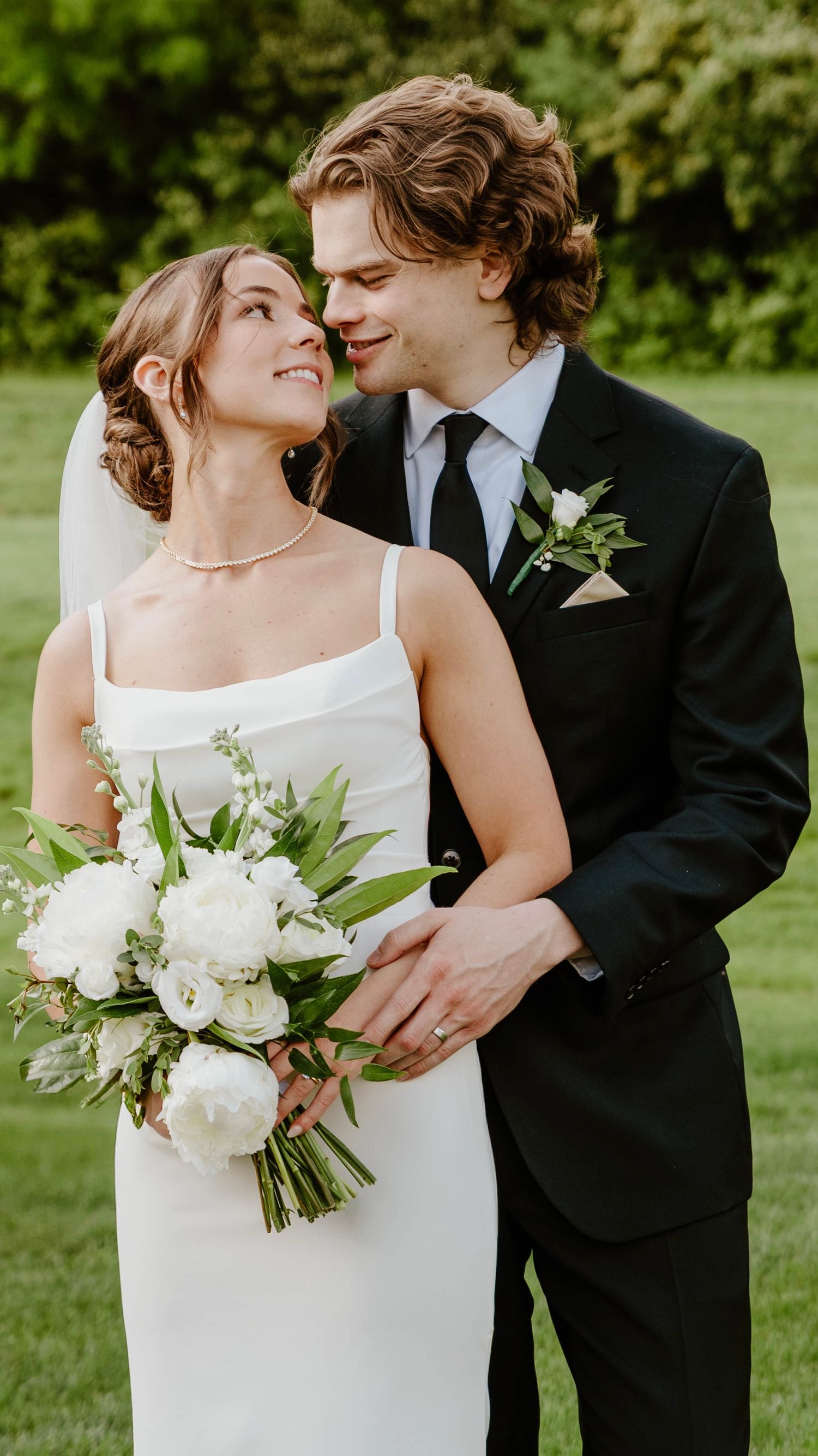 Romantic wedding couple embraces outdoors with white bouquet and classic black tuxedo against green garden backdrop.