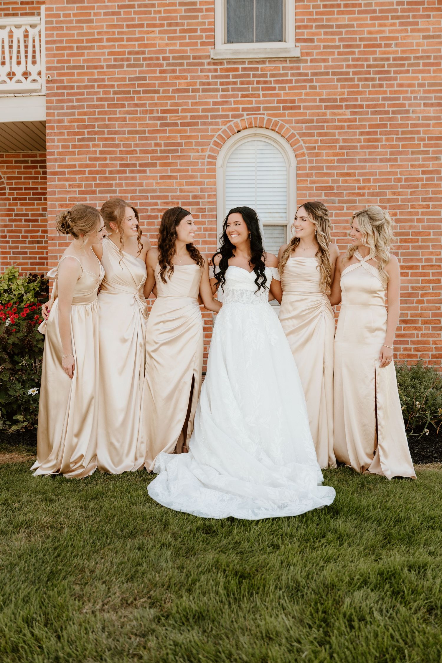 A bride in white and bridesmaids in champagne dresses pose together on grass against a brick church wall.