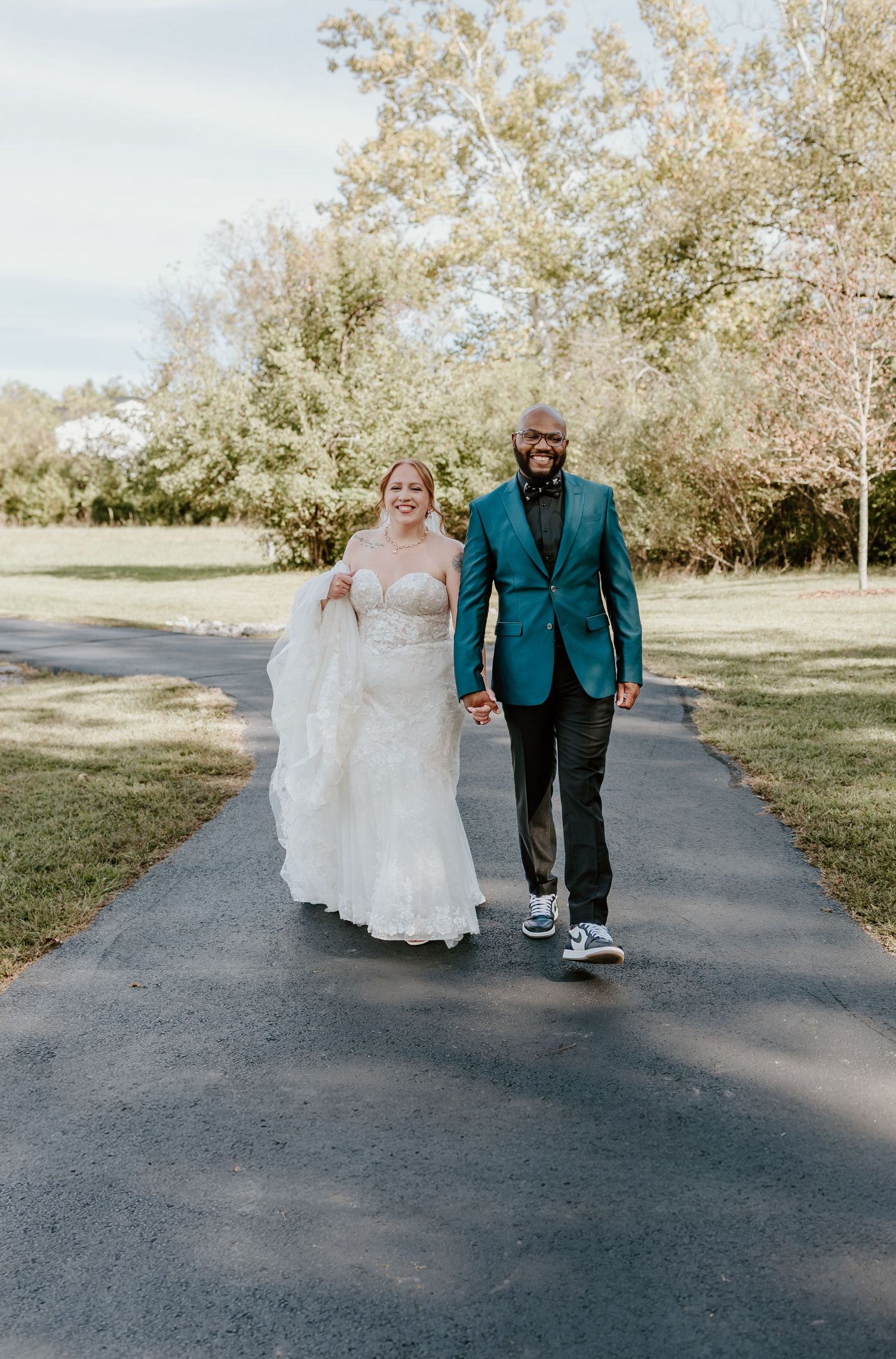 A couple walks down a tree-lined path, with the bride in a flowing white dress and groom in a teal blazer and sneakers.