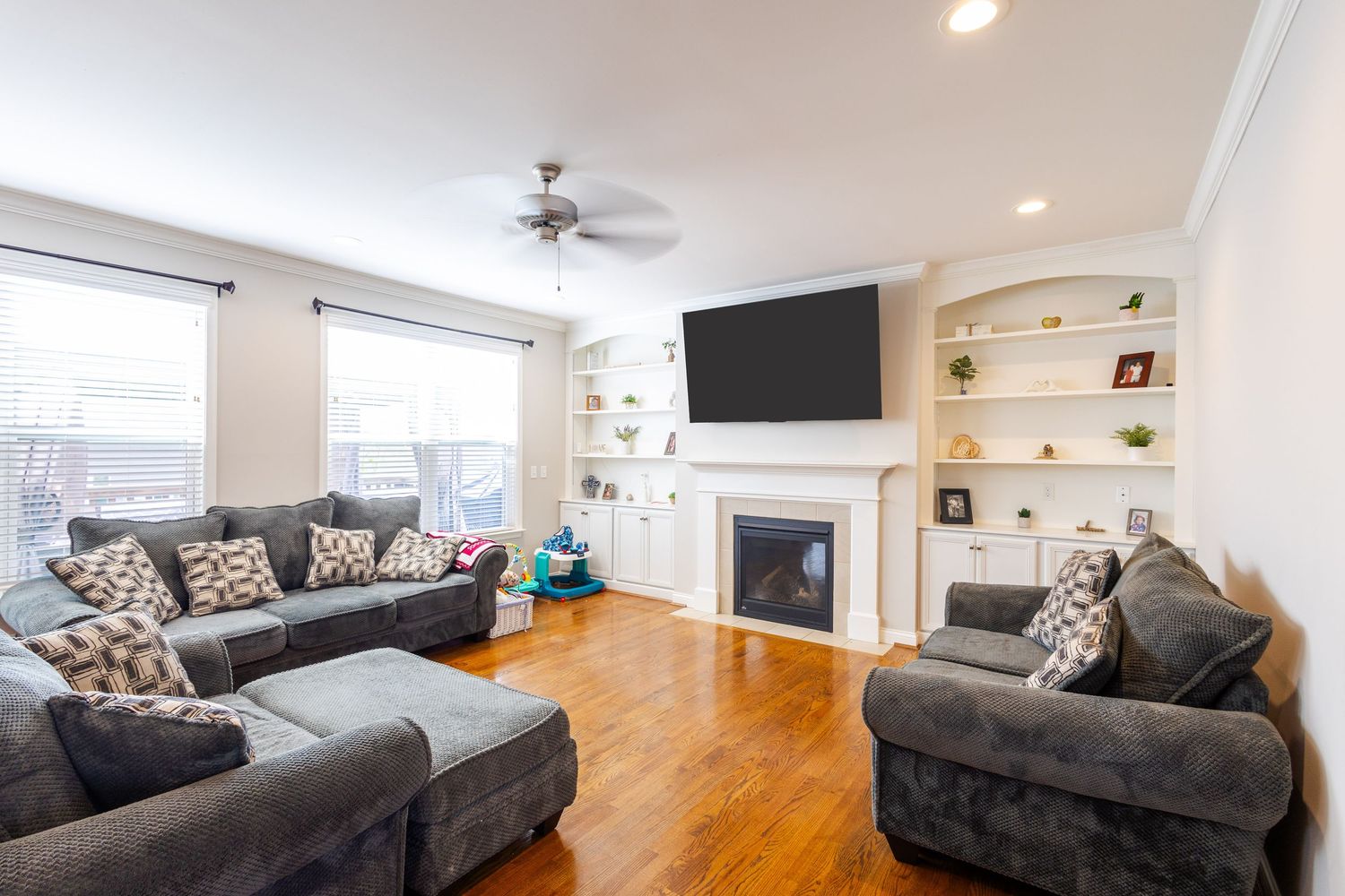 Cozy living room with gray sectional sofas, hardwood floors, built-in shelves, ceiling fan, and a fireplace with wall-mounted TV.