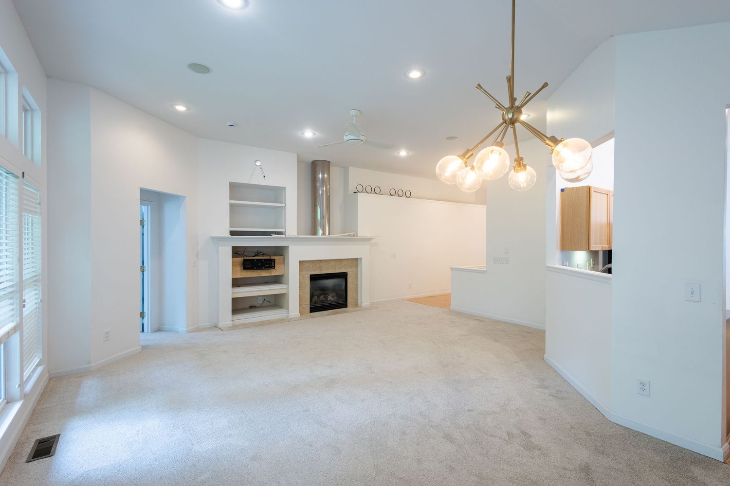 Bright open-concept living room with white walls, carpet, built-in shelves, fireplace, and modern gold chandelier.