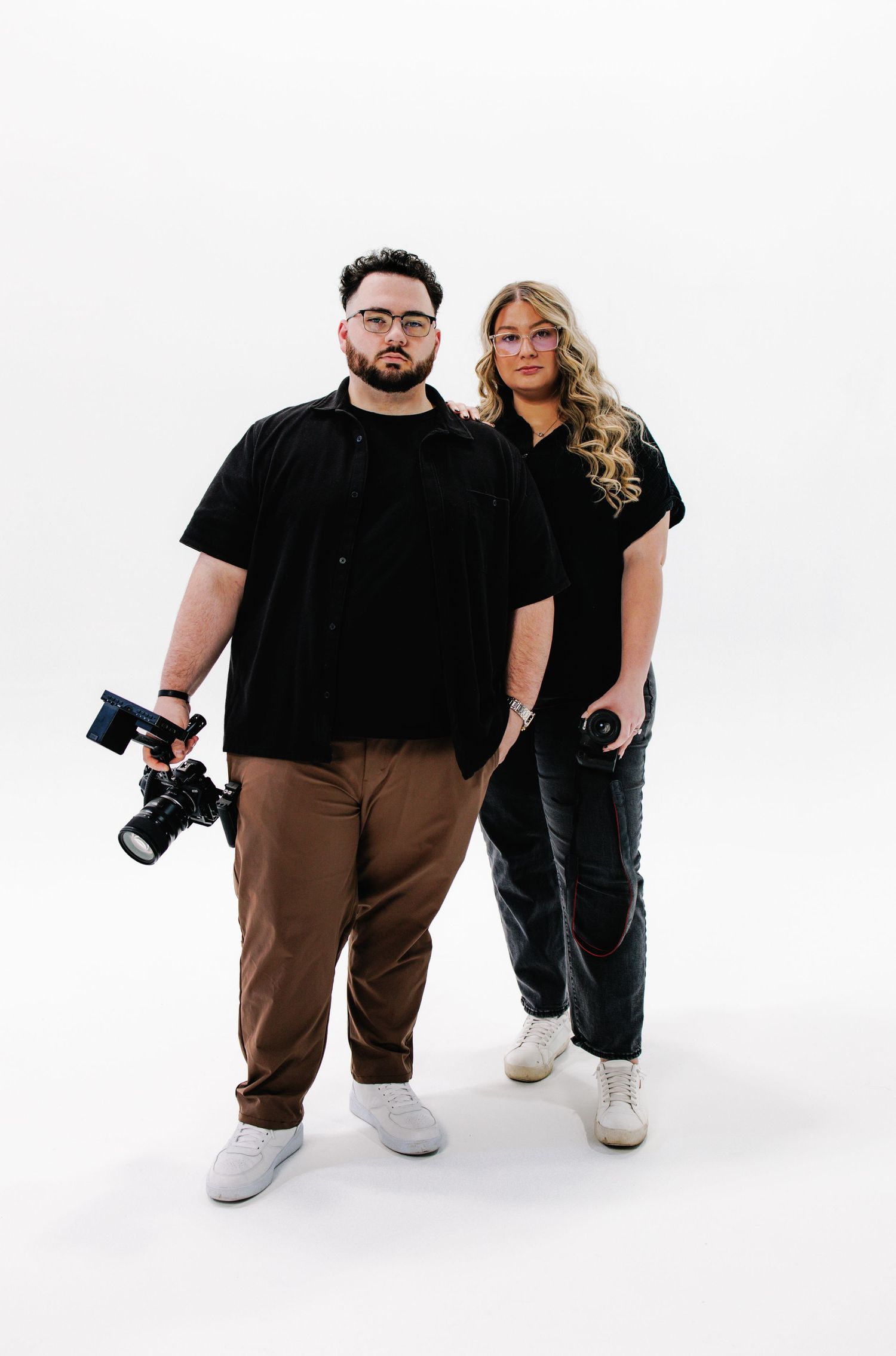 Two photographers in black and brown outfits pose with cameras against a white studio background.