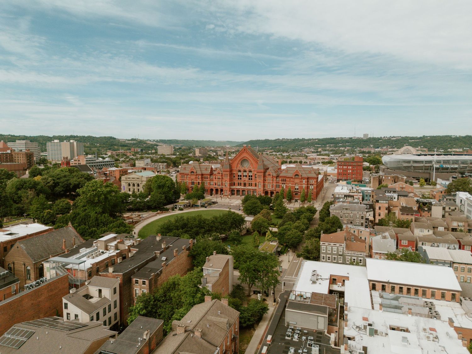 Aerial view of Cincinnati cityscape featuring historic red brick buildings, lush green trees, and a partly cloudy sky.