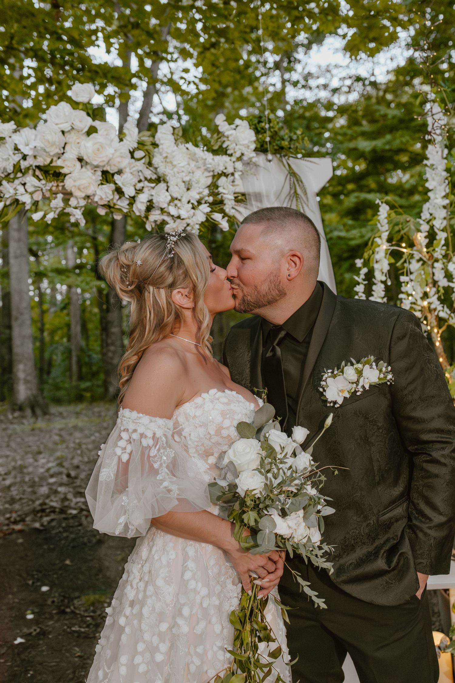 A romantic wedding couple shares tender kisses under hanging floral decorations in a woodland setting.
