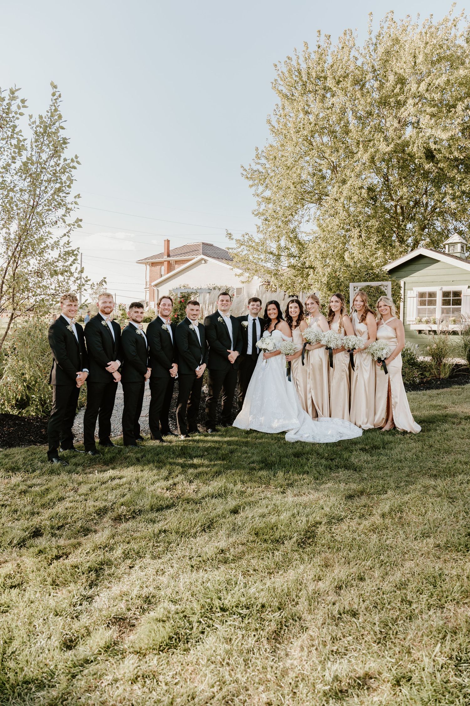 Wedding party poses outdoors on a grassy lawn with white and beige attire under blooming spring trees.