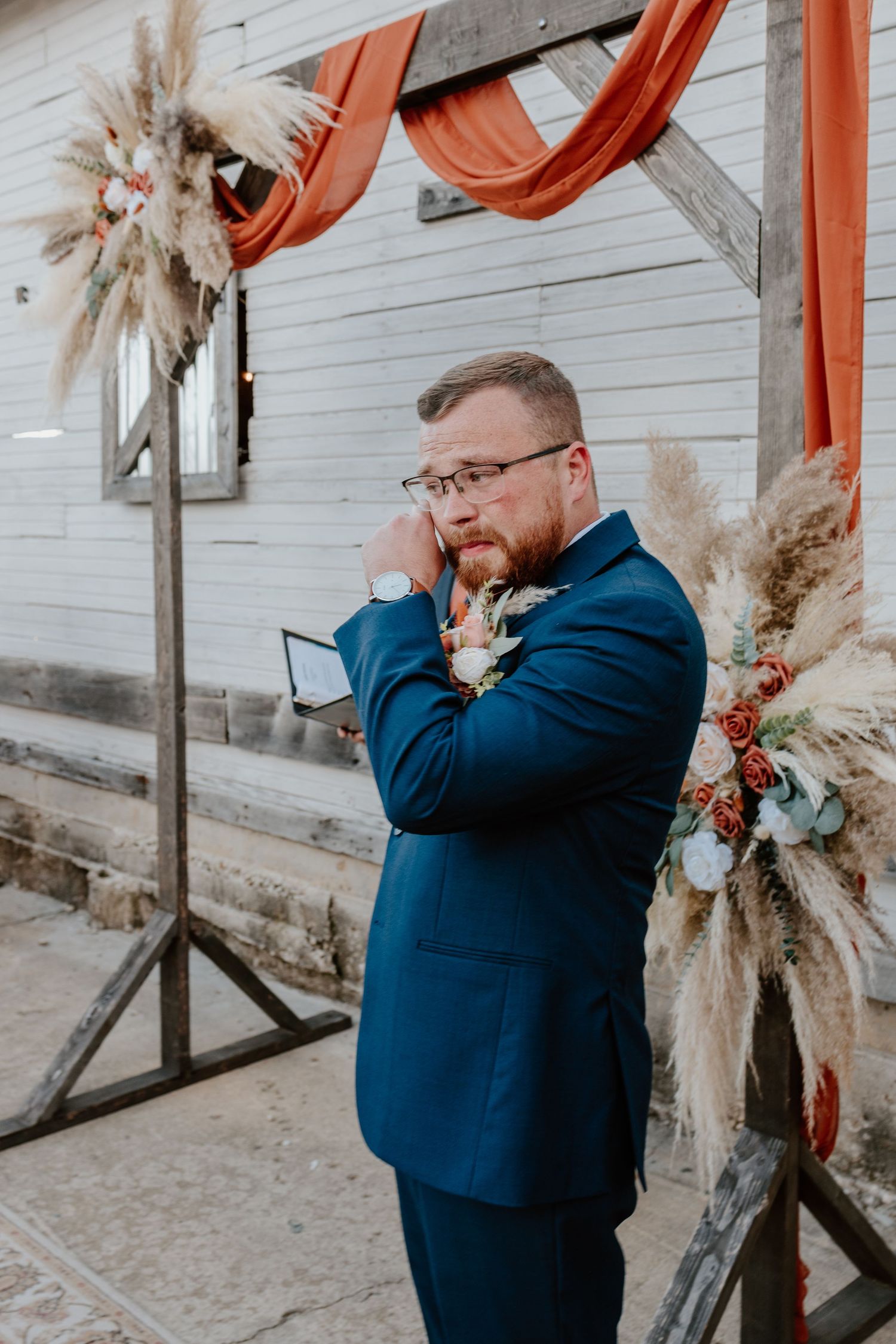 A person in a navy suit stands against a rustic backdrop with dried pampas grass and orange ribbon decor.