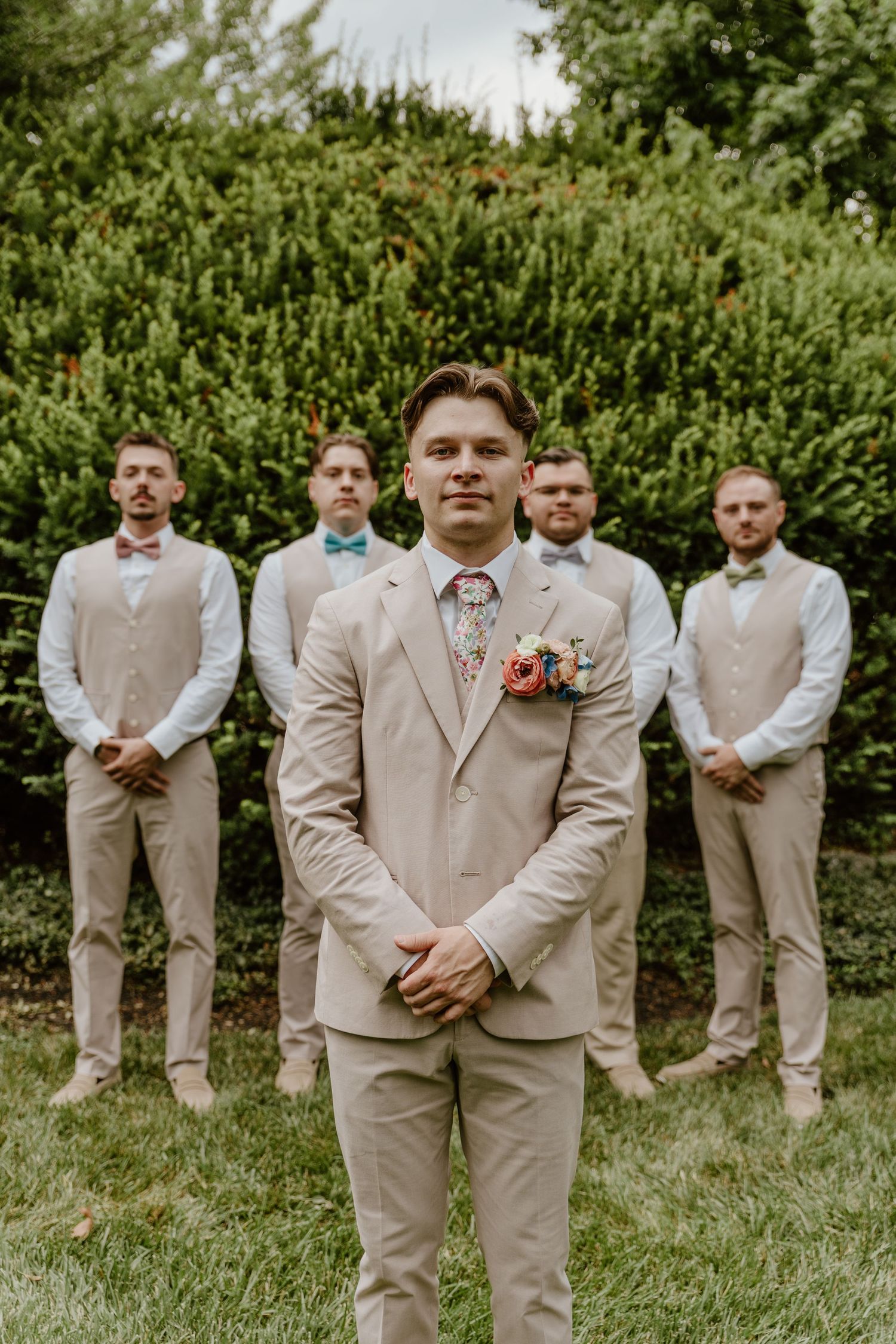 Groom in beige suit with floral tie and boutonniere stands before four groomsmen in matching vests and bow ties outdoors.