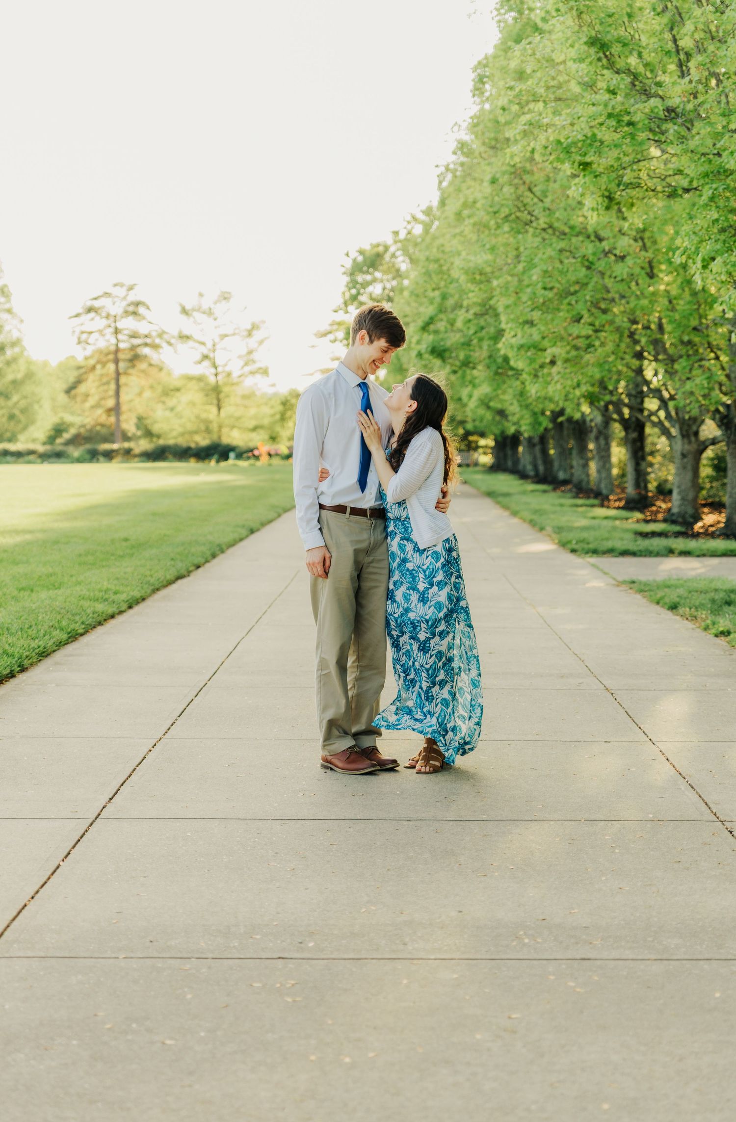 A couple stands together on a long concrete path surrounded by green trees.