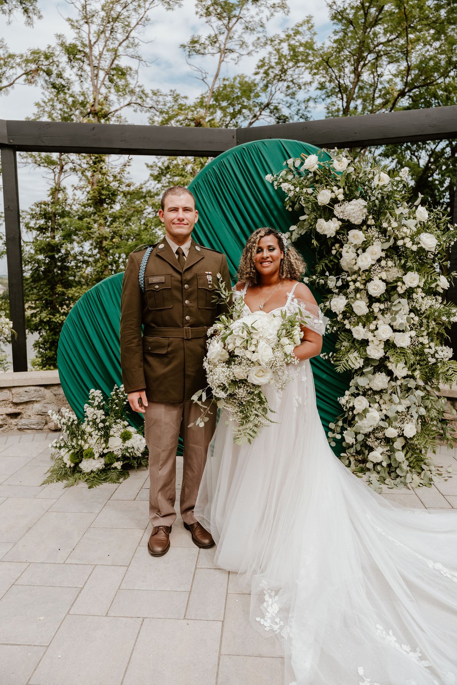 Bride and groom in military uniform pose before a green floral arch with white blooms at outdoor wedding ceremony.