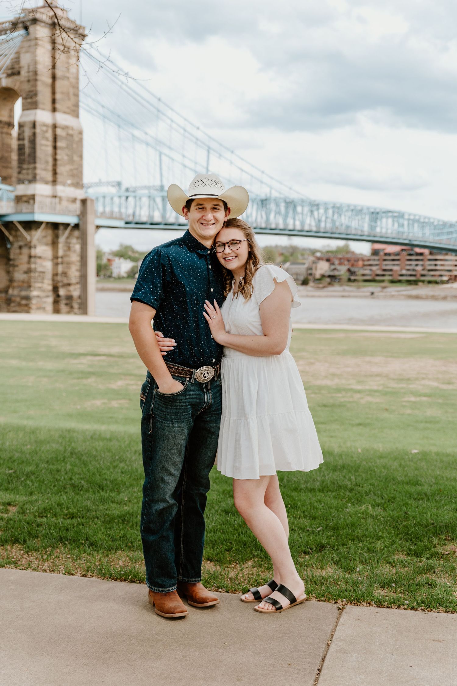 Couple in western attire poses near historic bridge with river and city skyline in background.