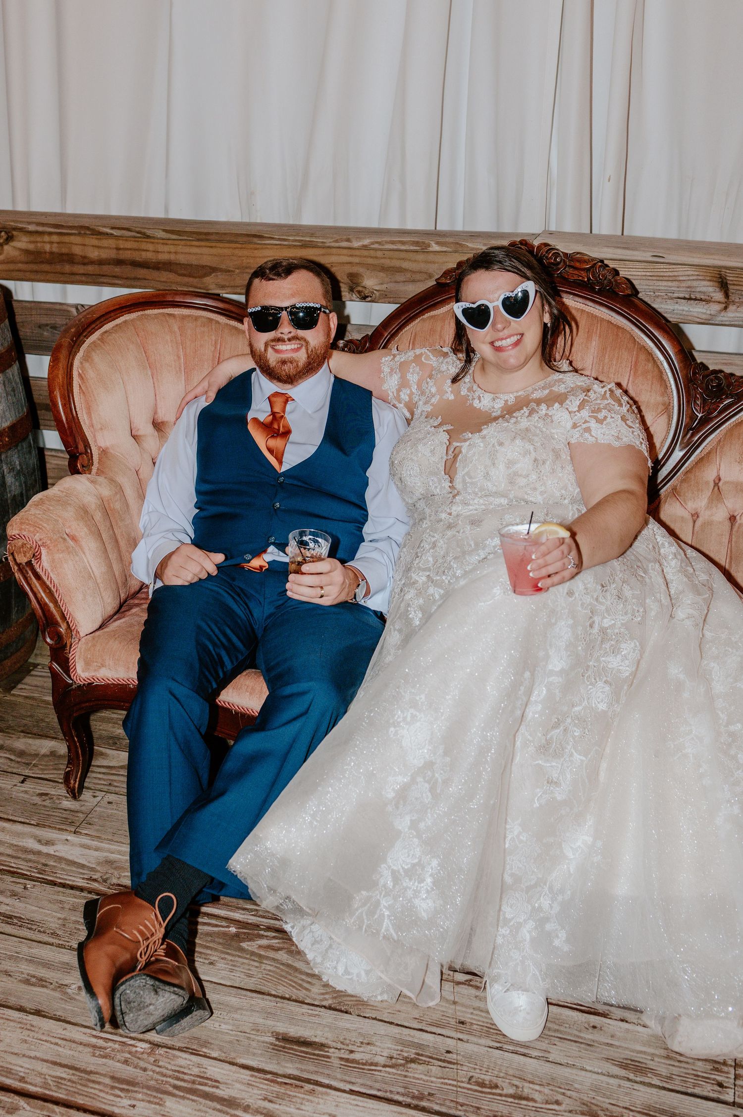 A couple in wedding attire sits on a vintage sofa wearing sunglasses while looking at their phones.