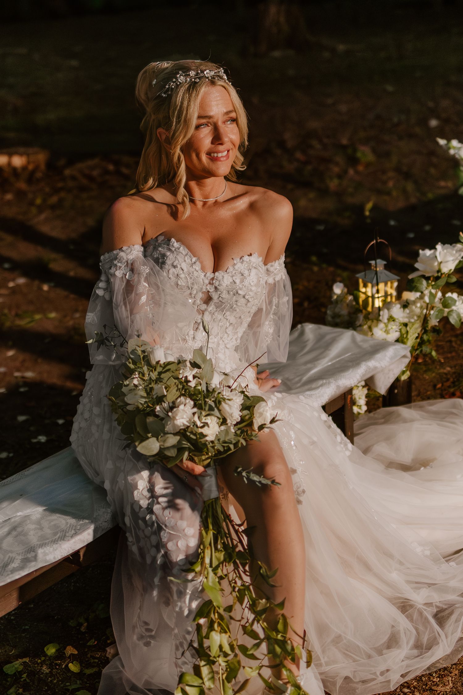 A bride in a flowing lace wedding dress poses in a natural outdoor setting with a white and green floral bouquet.