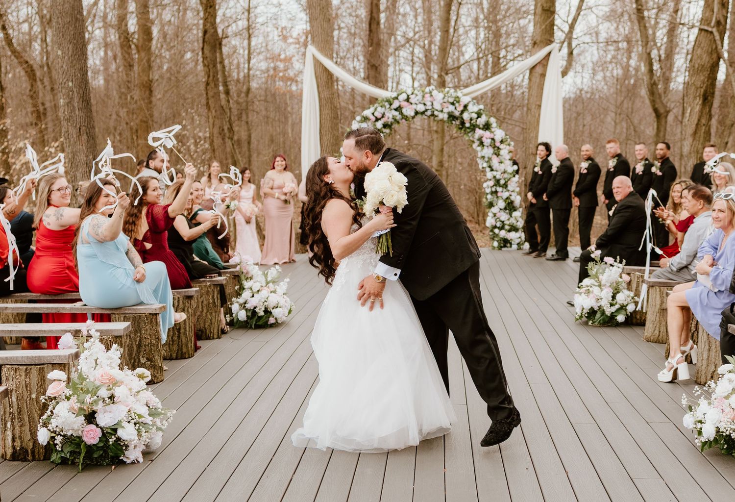 Bride and groom share a kiss at outdoor wedding ceremony on wooden deck with floral arch and guests cheering.