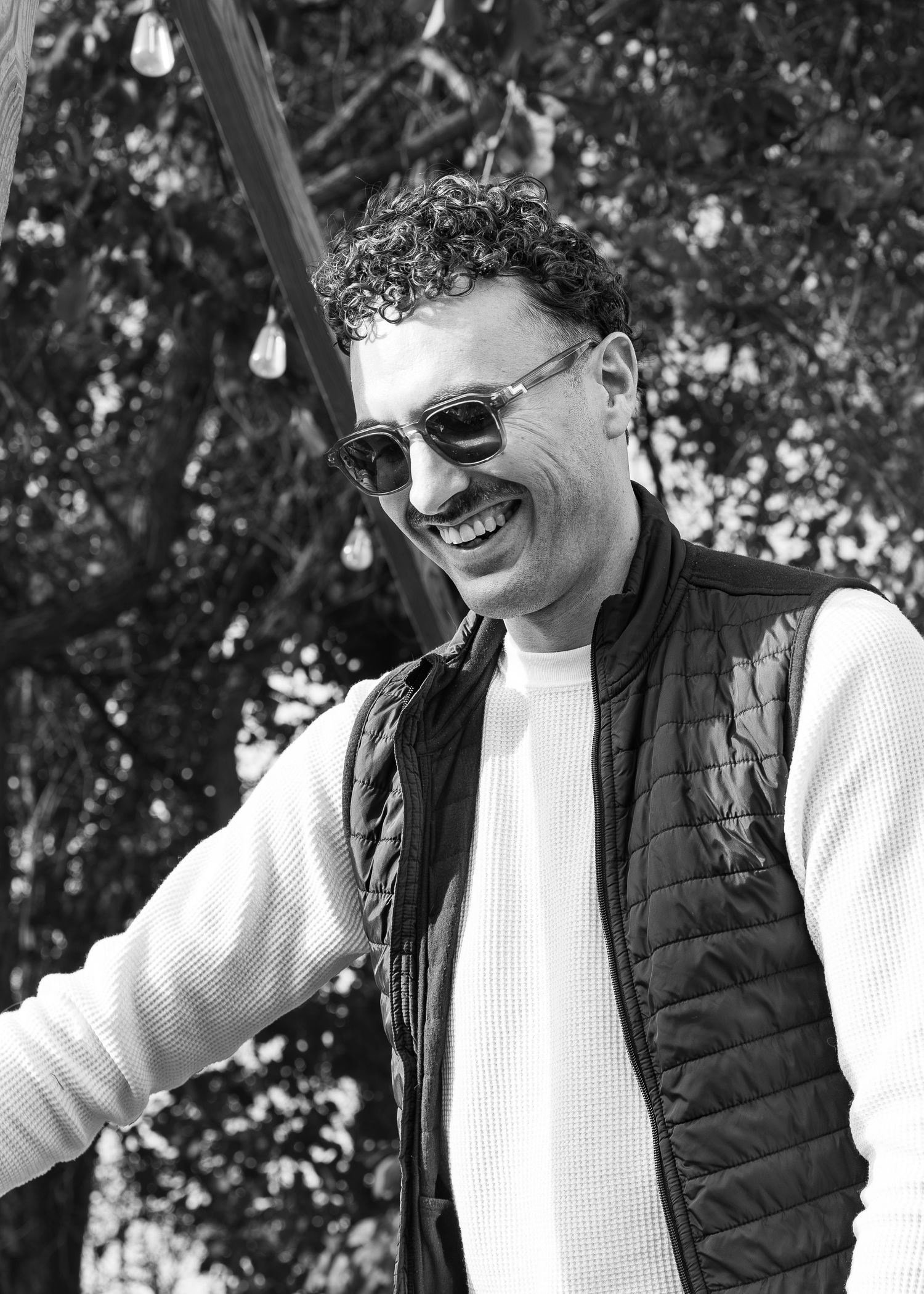 Person in black vest and white sweater smiling and gesturing while standing outdoors with string lights visible.